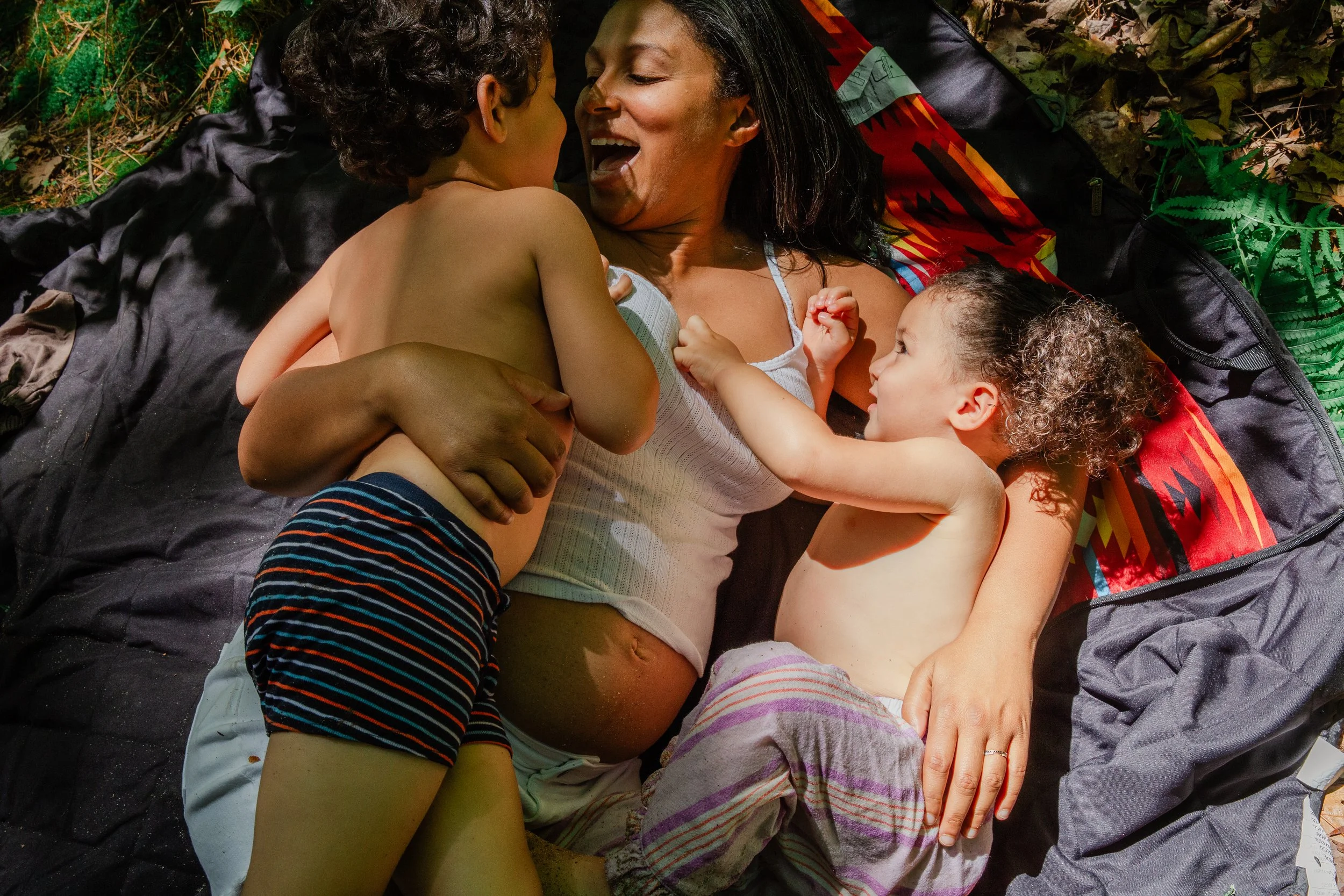 A woman lying on a blanket outdoors with three children, all smiling and cuddling, surrounded by leaves and grass.