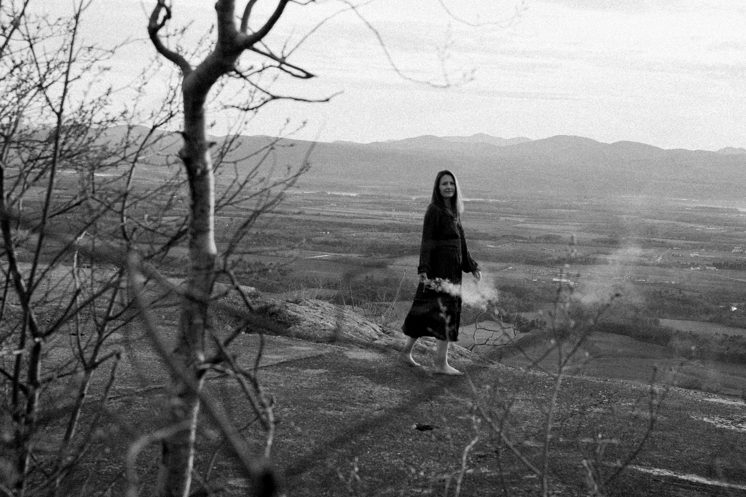 A woman in a skirt and jacket standing outdoors on a uneven terrain with a landscape of fields and mountains in the background. Dry branches are in the foreground, black and white photo.