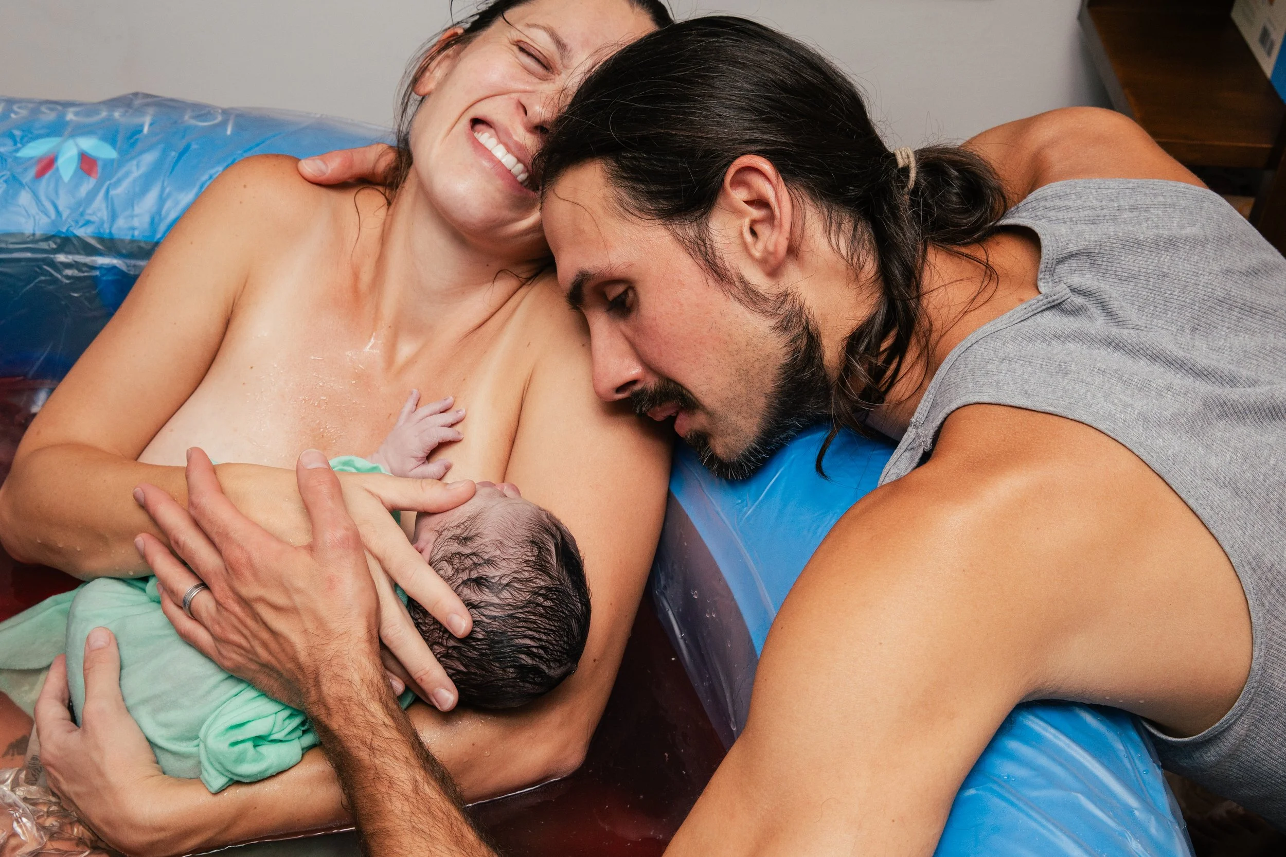 A woman and a man are holding a newborn baby, likely during a home birth, with the woman smiling and the man kissing the baby's head.