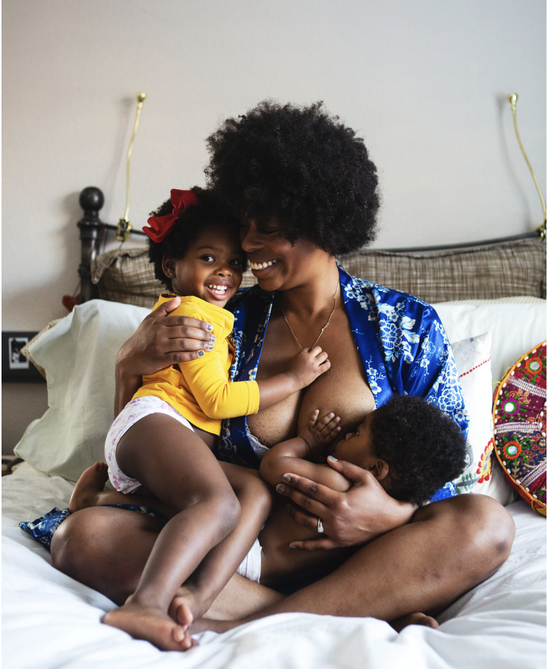 A woman with two children, one on her lap and the other hugging her, sitting on a bed in a cozy room. The woman is smiling while the children are happy.