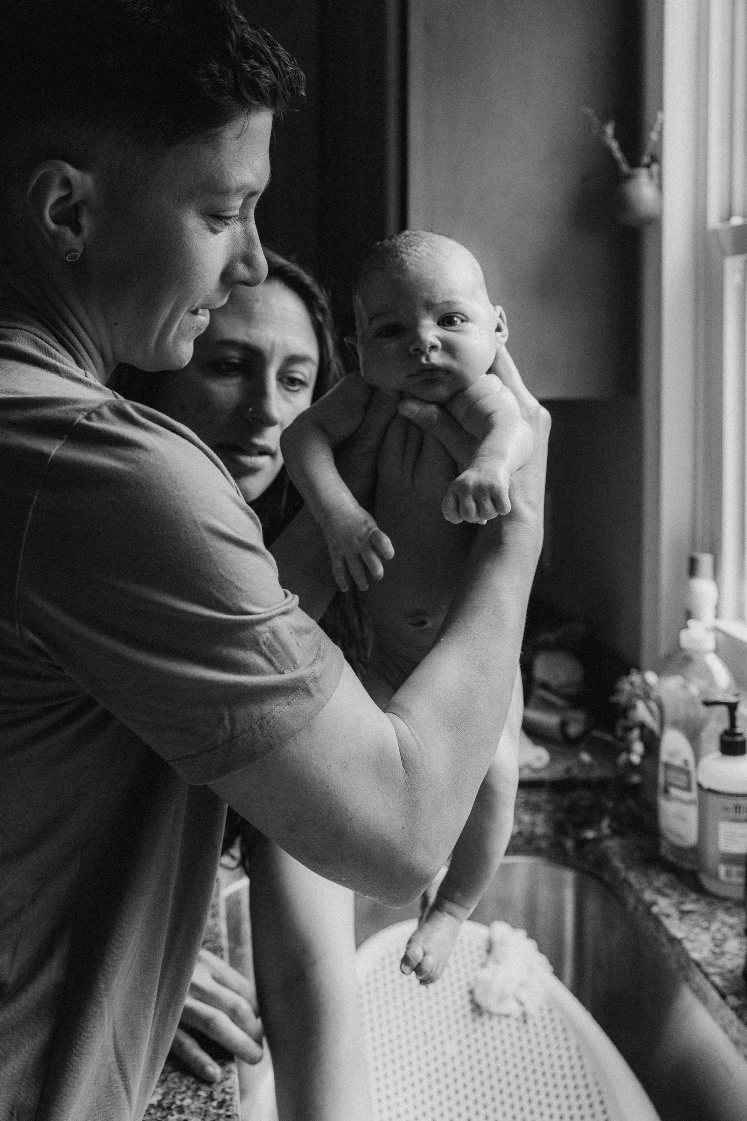A person holding a newborn baby in a kitchen, with a woman standing next to them, looking at the baby. The scene appears to be joyful and intimate, in black and white.