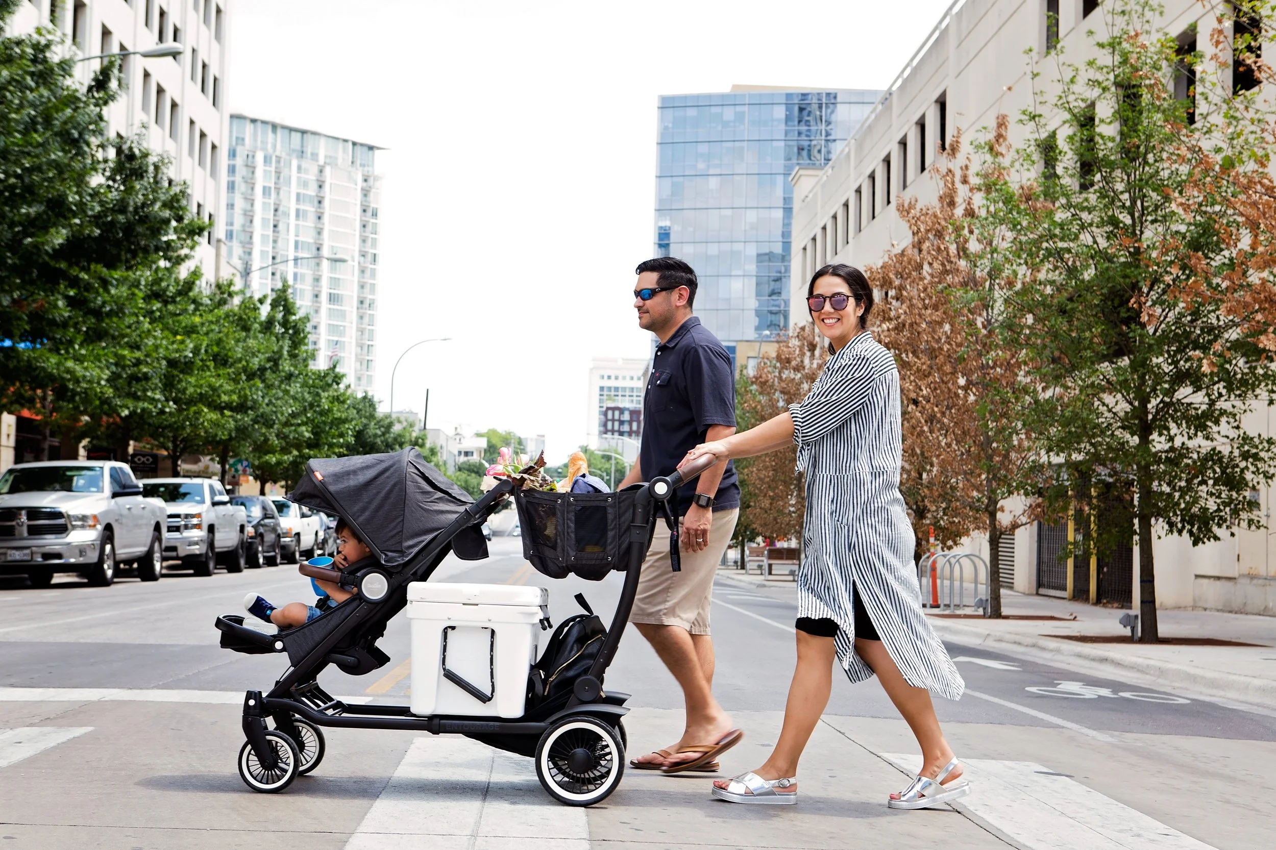 A family walking across a city street with their baby in a stroller, with tall buildings and green trees in the background.