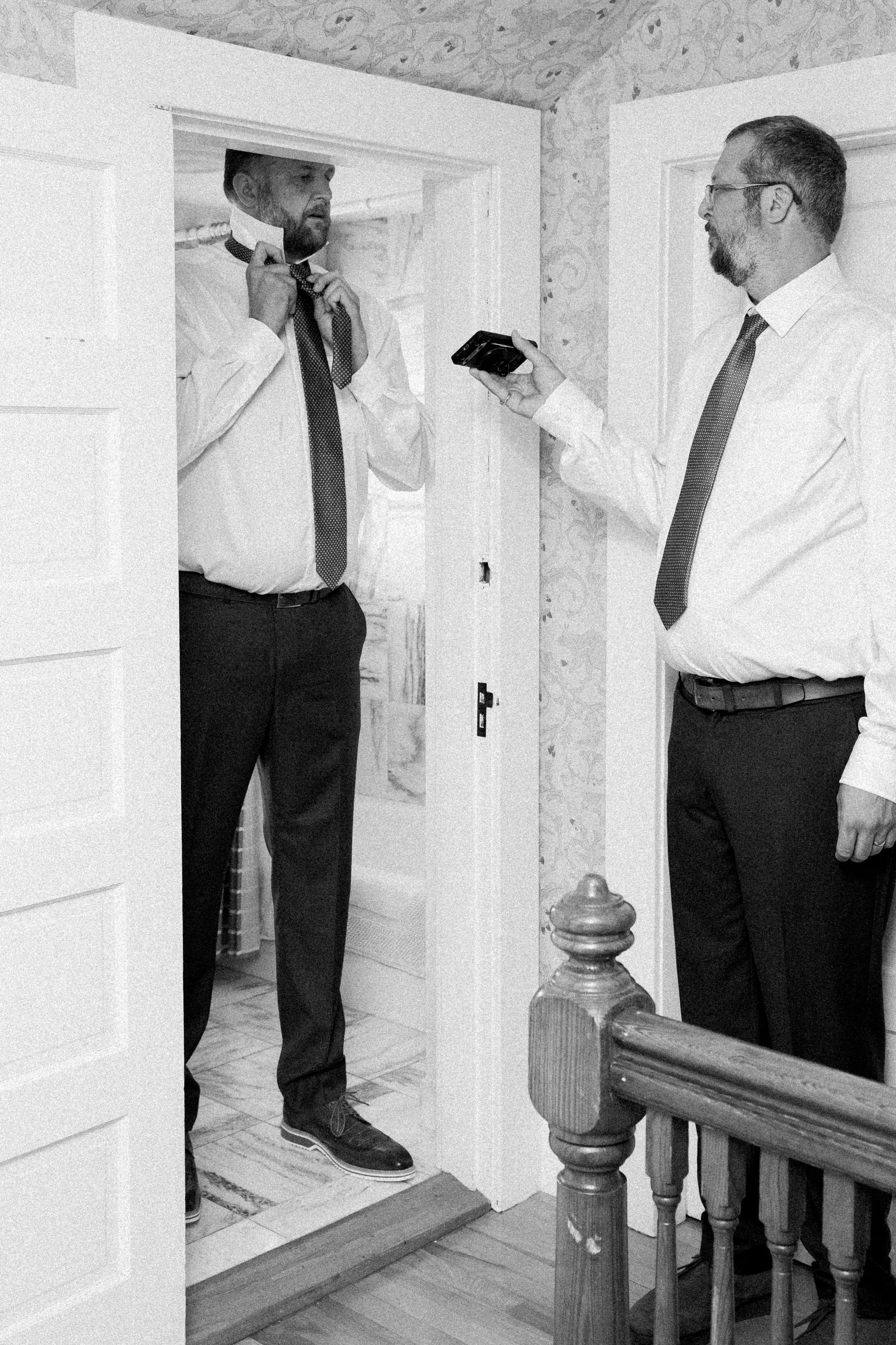 Two men in office attire standing in a doorway, with one adjusting his tie and the other holding a phone, inside a home with patterned wallpaper and wooden staircase.