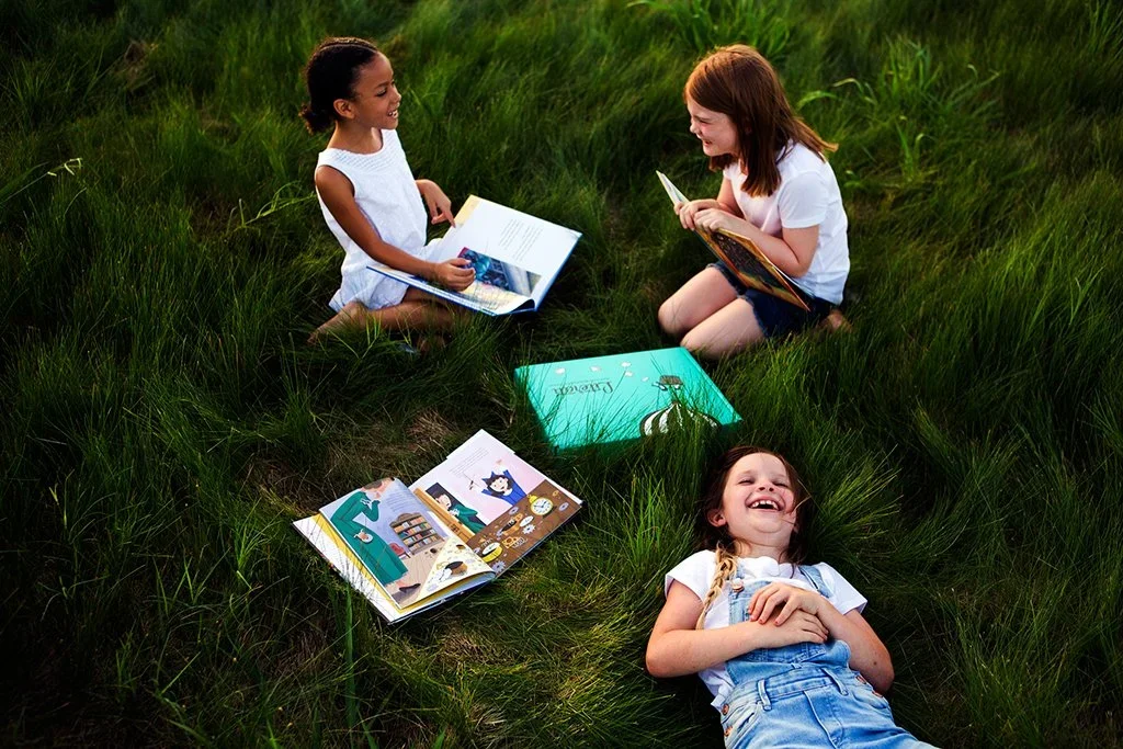 Three young girls sitting and lying in tall grass, reading and looking at picture books, outdoors on a grassy field.