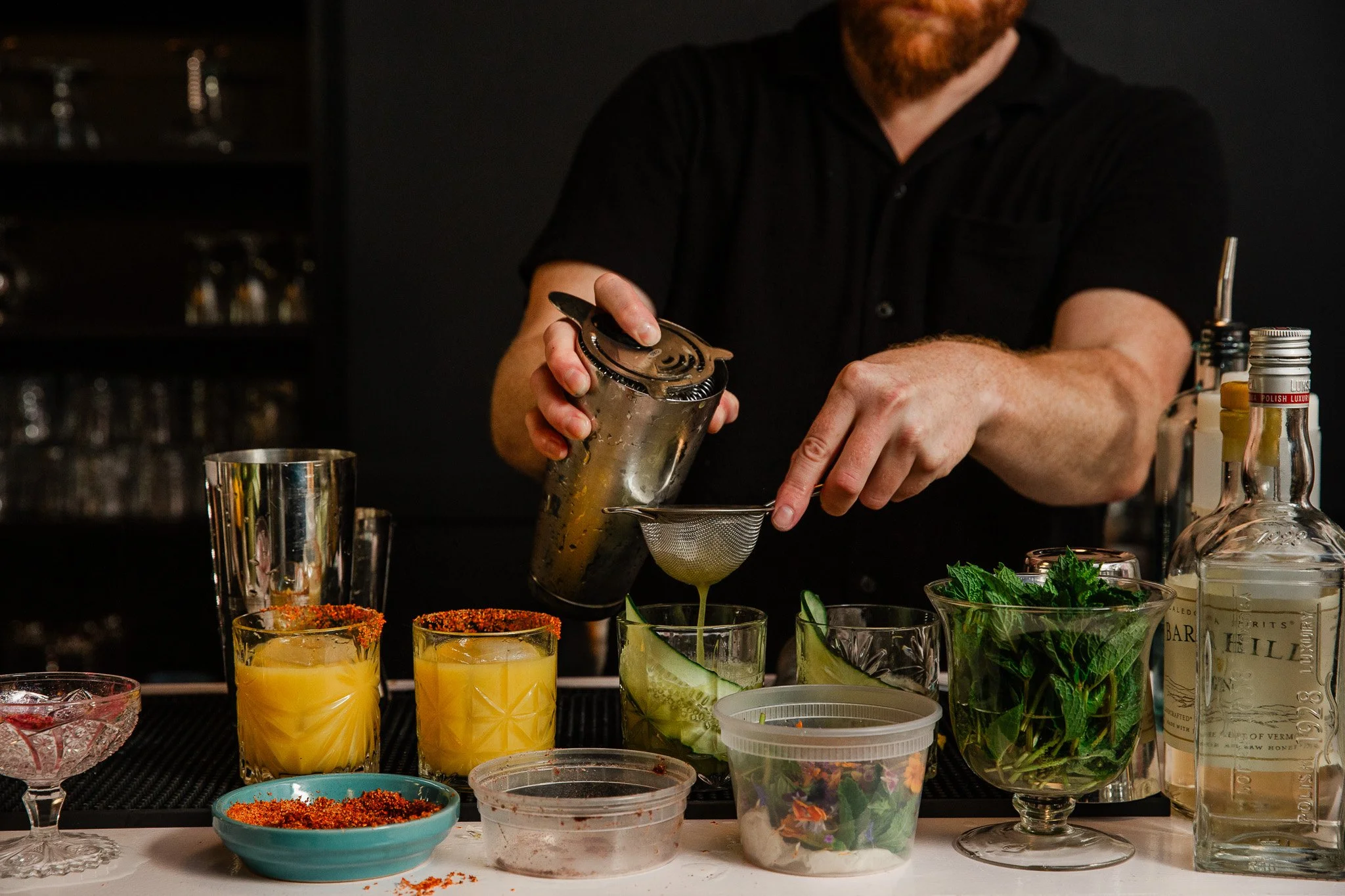 A bartender pouring a yellow cocktail through a fine strainer into glasses garnished with lime and mint, with various bottles, herbs, and bar tools on the counter.