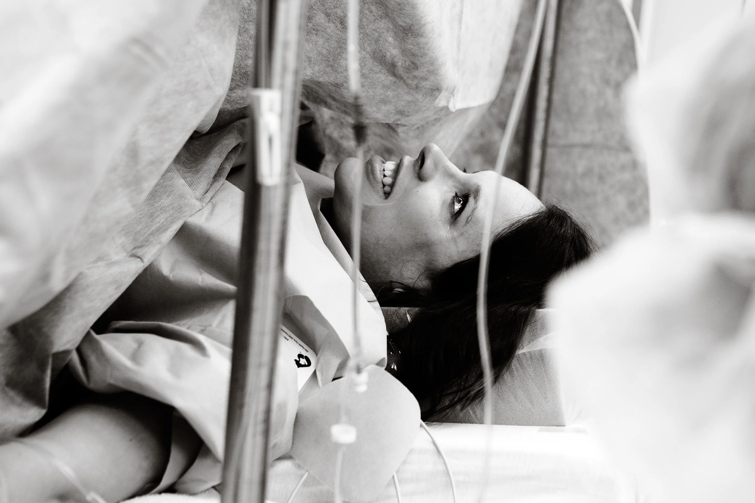 A woman smiling while lying in a hospital bed, viewed from underneath surgical railing, in black and white.