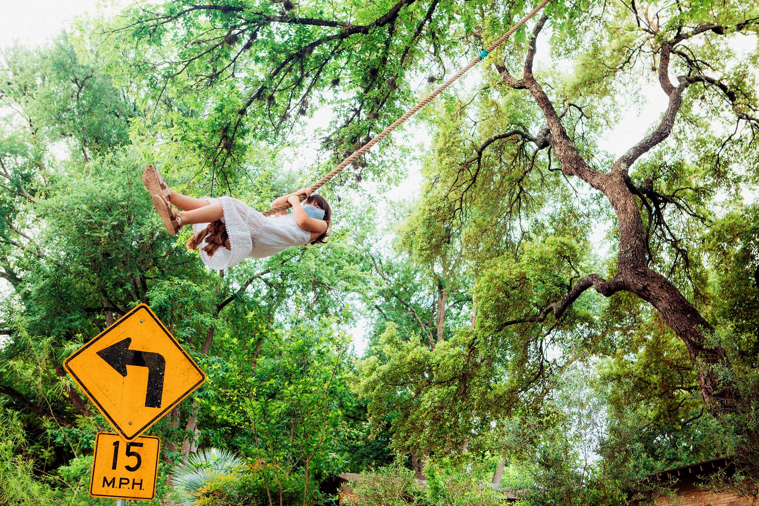 A young girl wearing a white dress and face mask, sitting on a rope swing hanging from tree branches in a lush green forest.