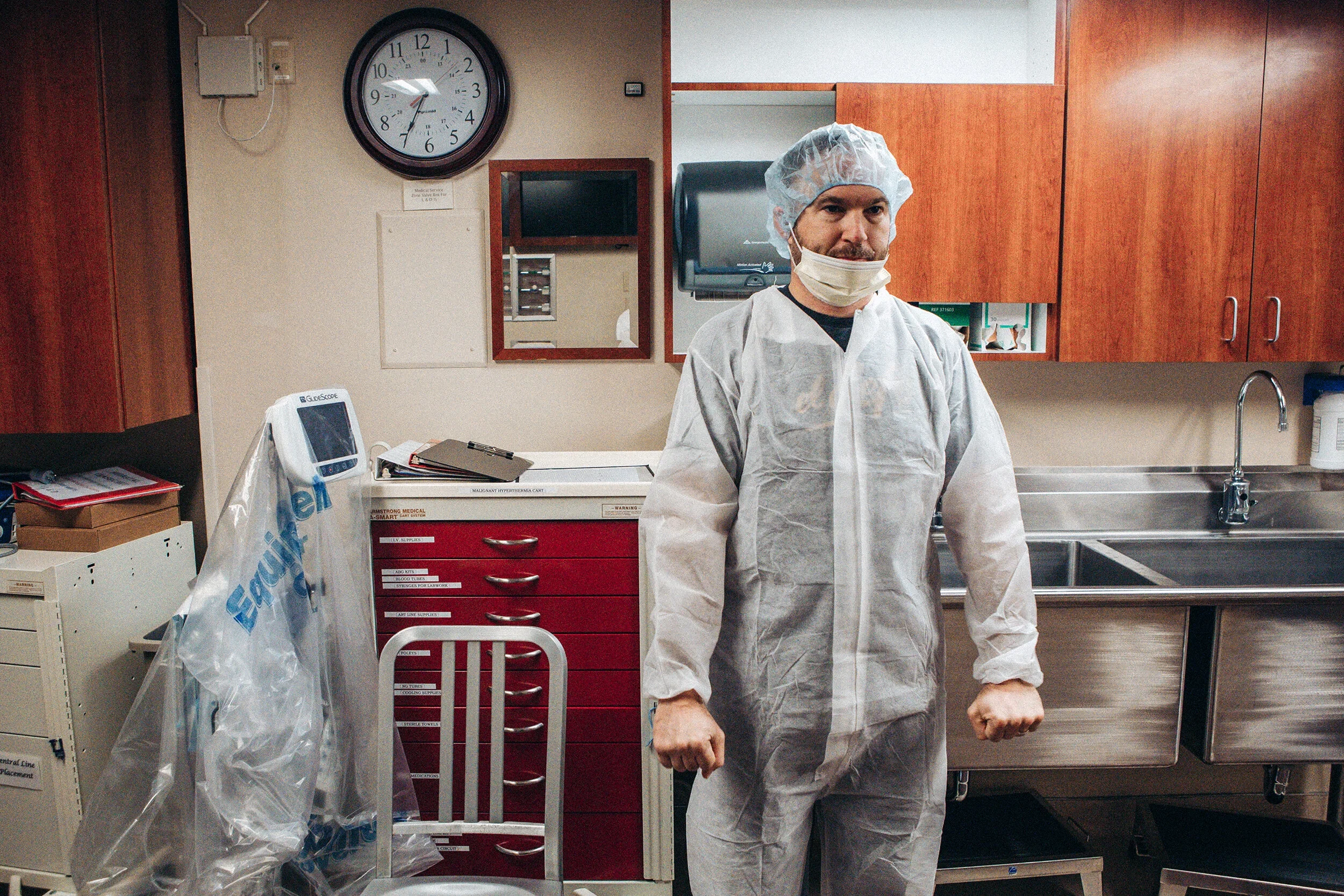 A male healthcare worker wearing a white protective gown, face mask, and hair cap standing in a hospital or clinical setting.