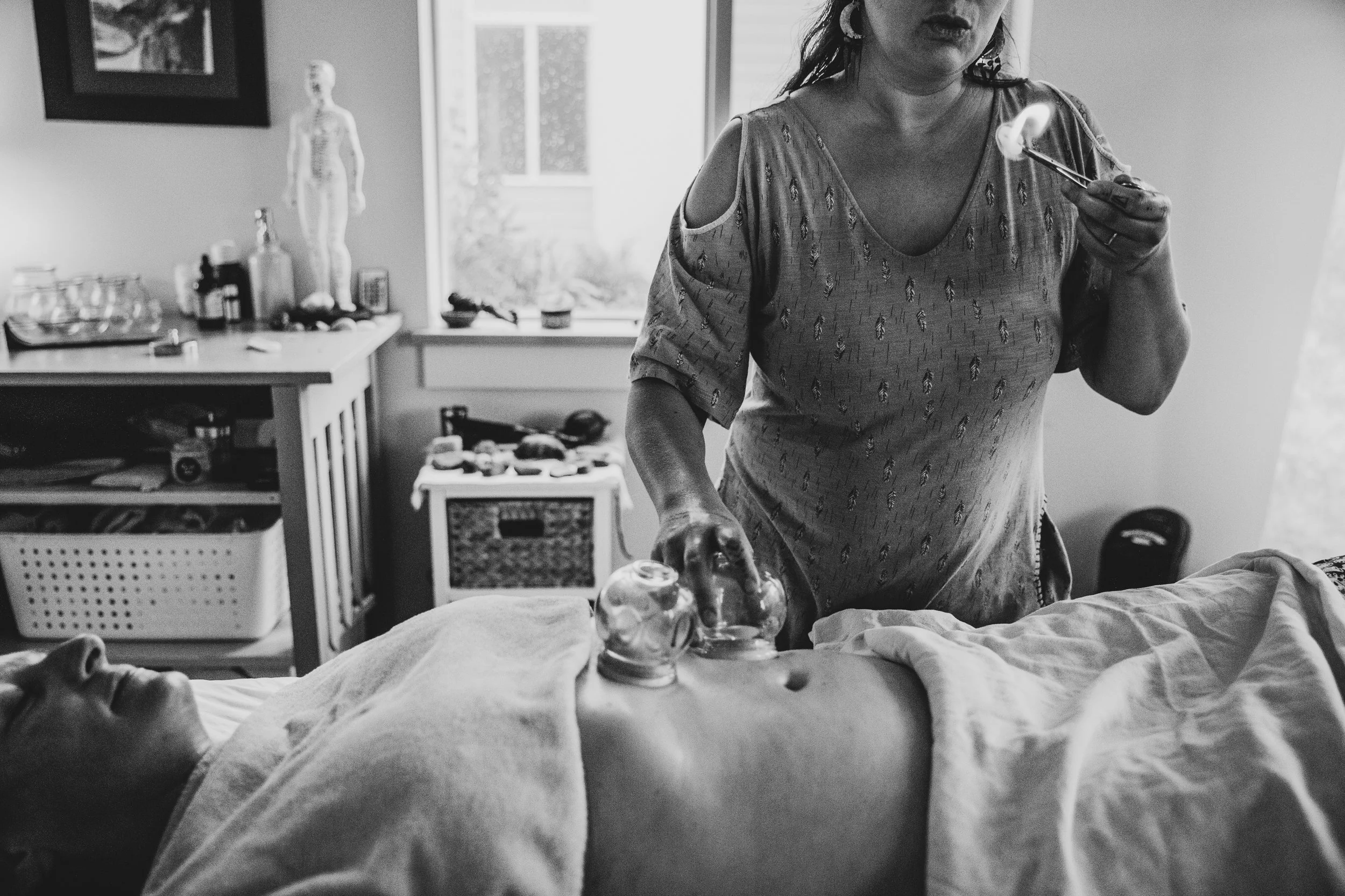 Black and white photo of a woman in a patterned shirt holding a spoon, standing next to a person lying on a bed, with medical supplies and a model of a human body in the background.