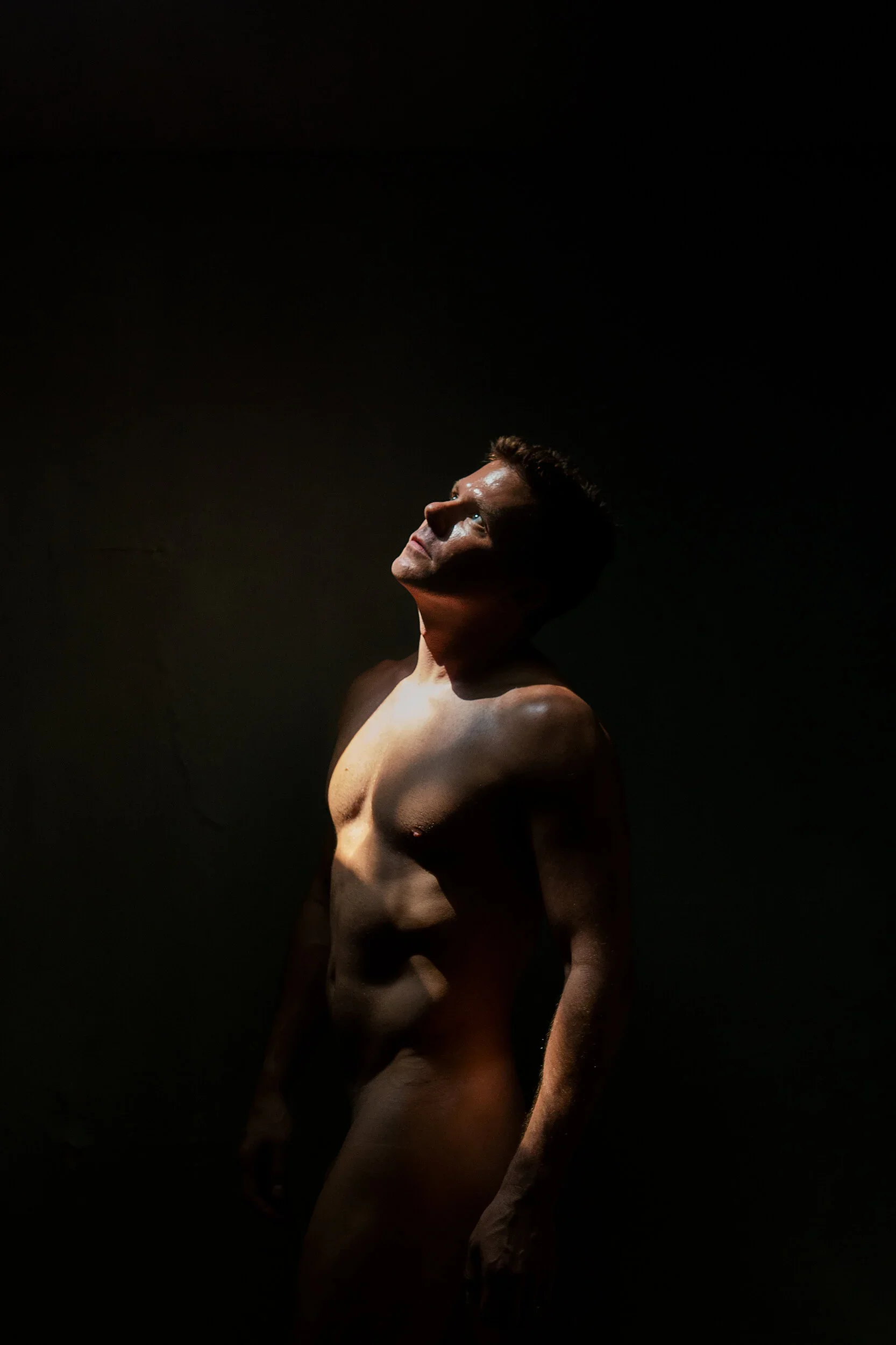 A shirtless man with short hair looking upward, illuminated by a focused light in a dark environment.