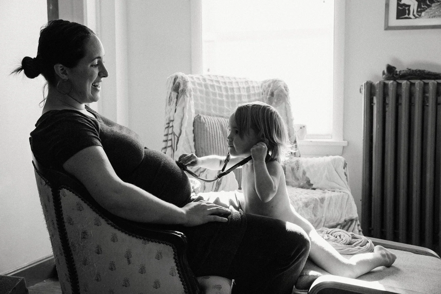 A woman sitting on a vintage armchair with a pregnant belly, smiling at a young child who is playing with a stethoscope. The child is sitting on an ottoman and touching the woman's stomach. The scene is in a well-lit room with a bed, drapes, and a ra