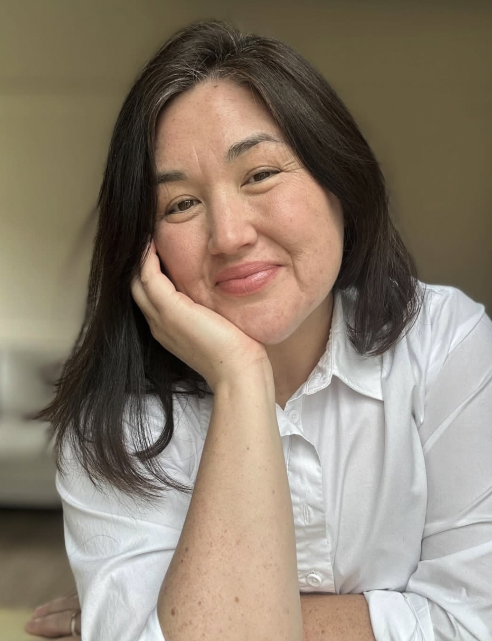 A woman with medium-length dark brown hair, smiling and resting her face on her hand, wearing a white button-up shirt, in a room with a yellowish wall background.