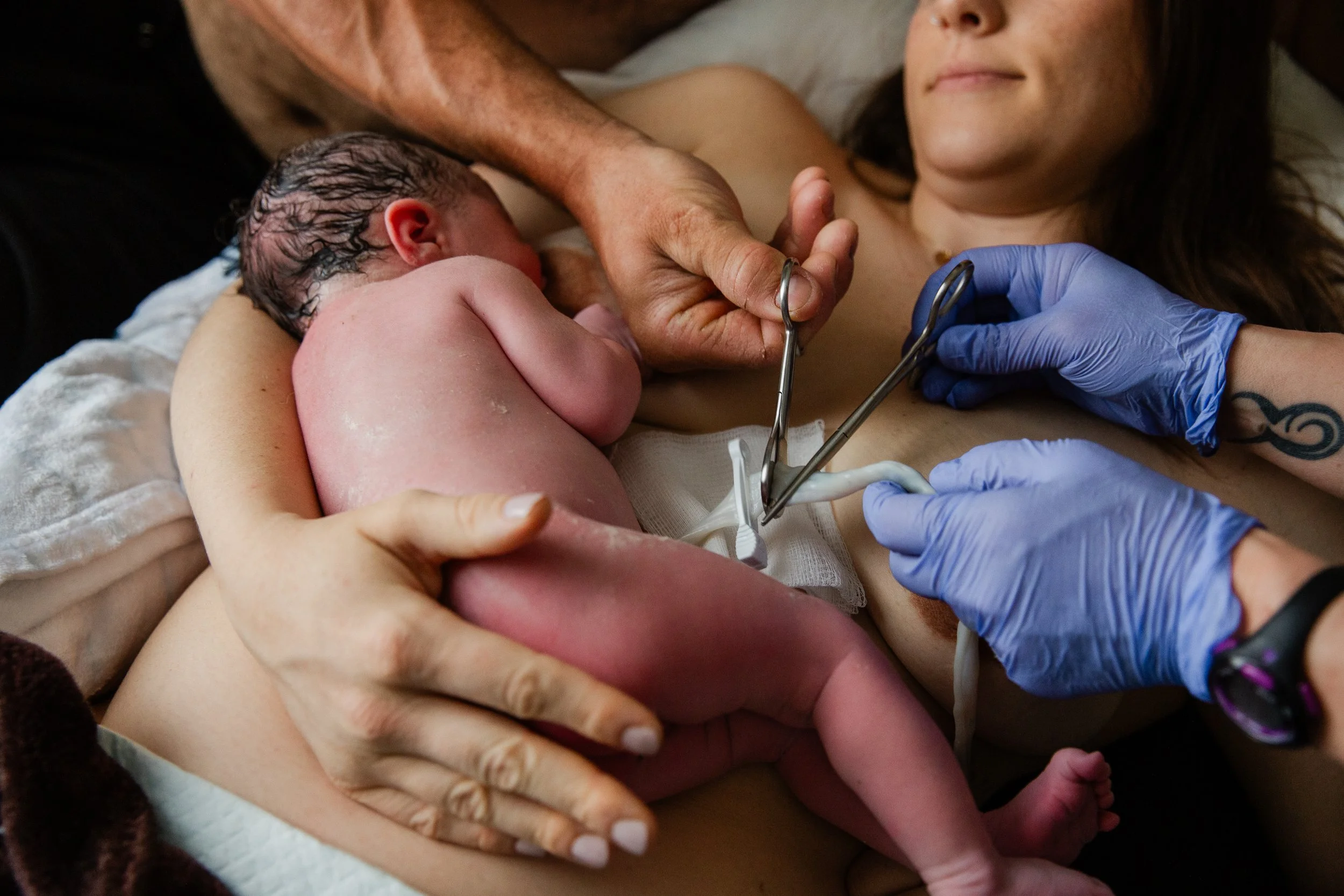 A newborn baby lying on a woman's chest during skin-to-skin contact, with a healthcare worker in blue gloves gently holding the baby's foot.
