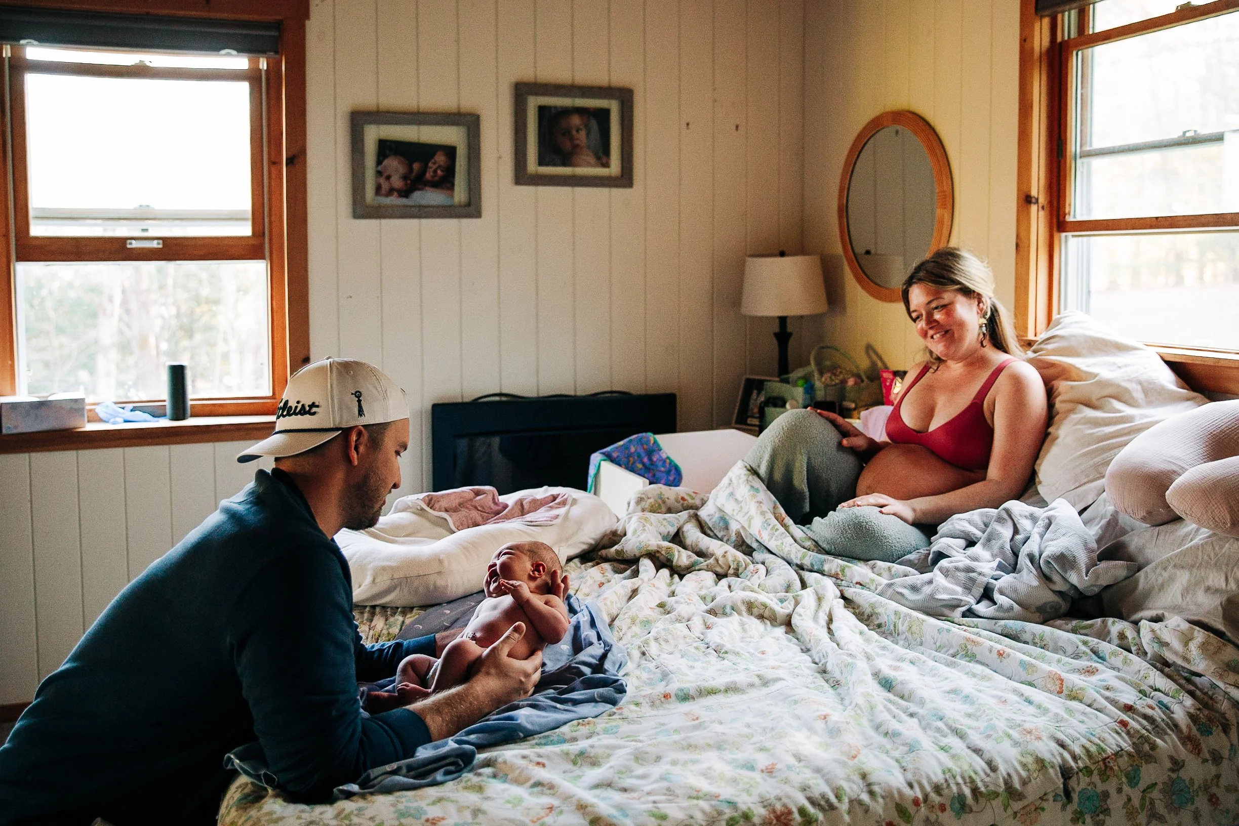 A woman sitting on a bed smiling at a baby held by a man, in a cozy room with natural light from windows, family photos on the wall, and a lamp beside the bed.