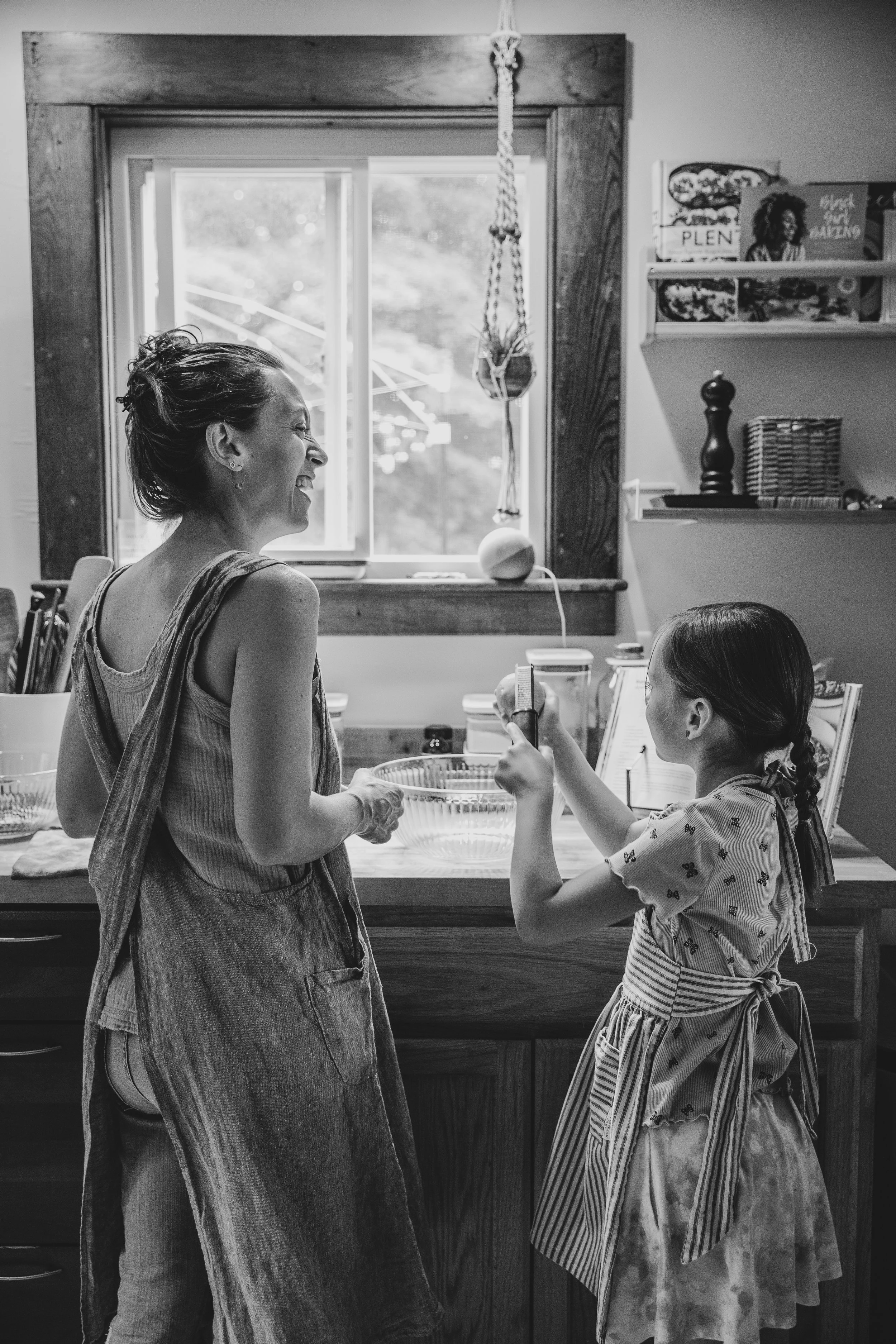 A woman and a young girl are in a kitchen, smiling and talking to each other. The woman has short hair and is wearing a sleeveless top and apron, holding a glass bowl. The girl has long hair in a braid, wearing a patterned dress with an apron, holdin