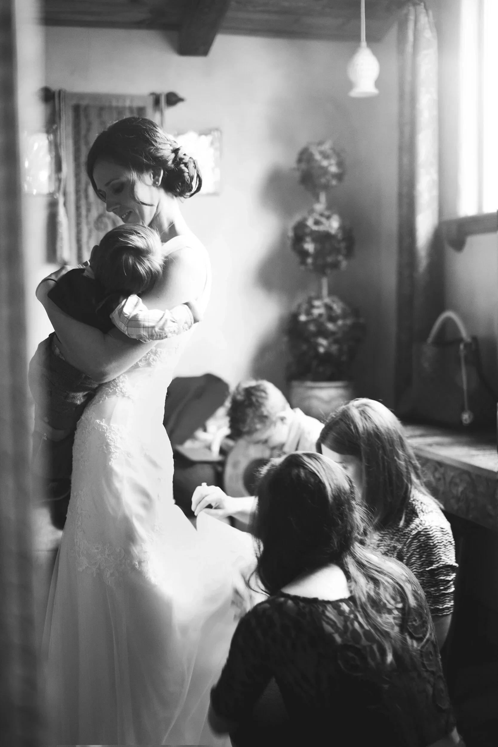A bride holding a young boy, smiling, while women sit and work on wedding dress details, in a warmly lit room.