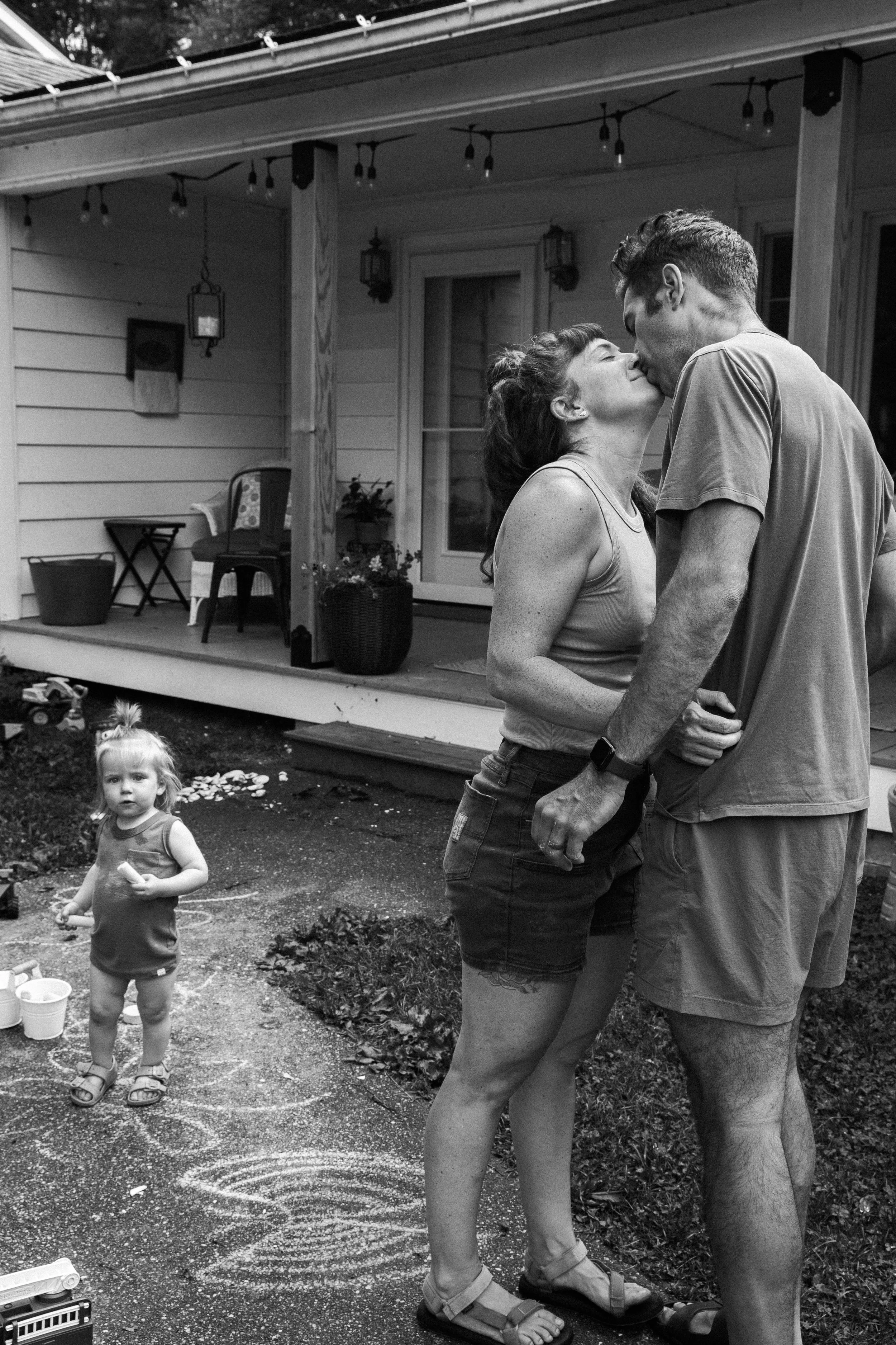 A couple is kissing on a porch in front of their house, while two young children are playing outside nearby, one girl looking at the camera and another child climbing a slide.