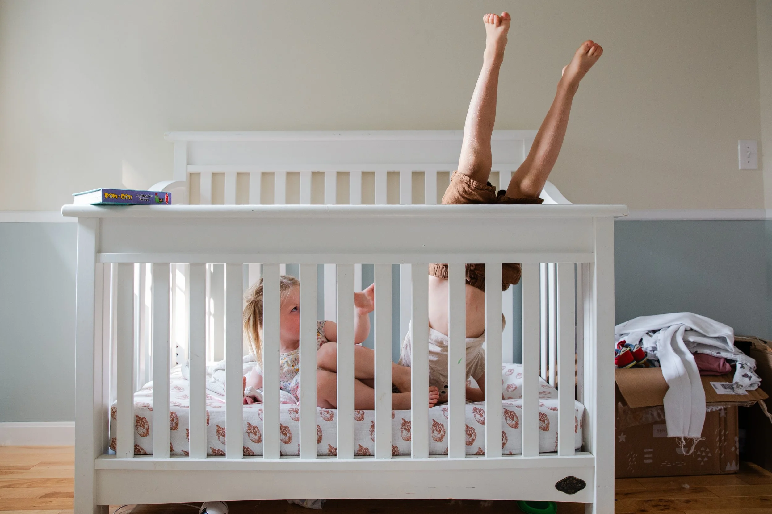 Childs legs sticking up above a white crib, with children sitting on a bed inside the crib looking up at the legs. There are books on top of the crib and miscellaneous items and boxes around it.