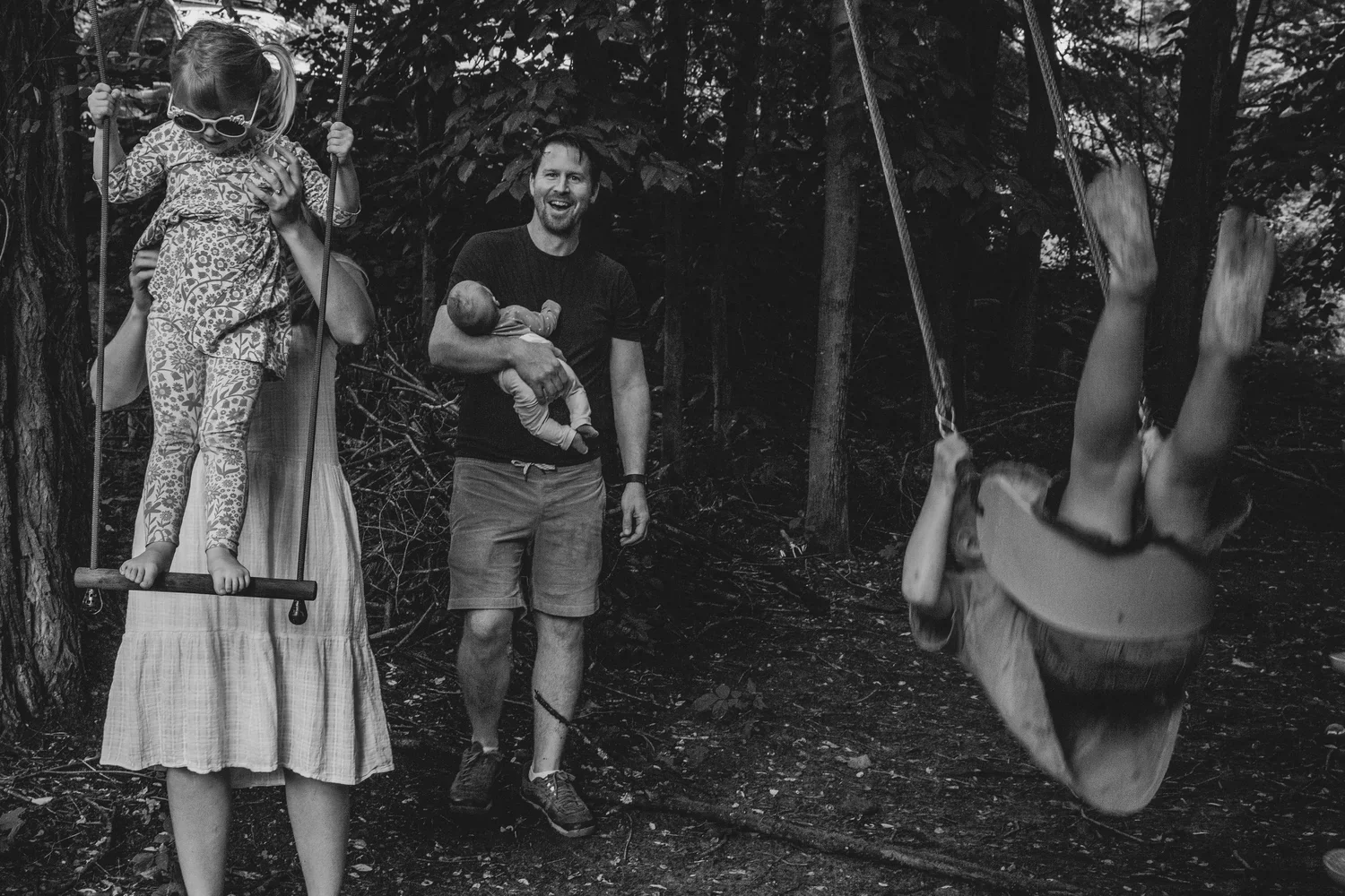 A woman and two children around a swing set in a wooded backyard. One girl in sunglasses is on a swing, an adult holding her, and a boy is mid-air on another swing, flipping upside down.