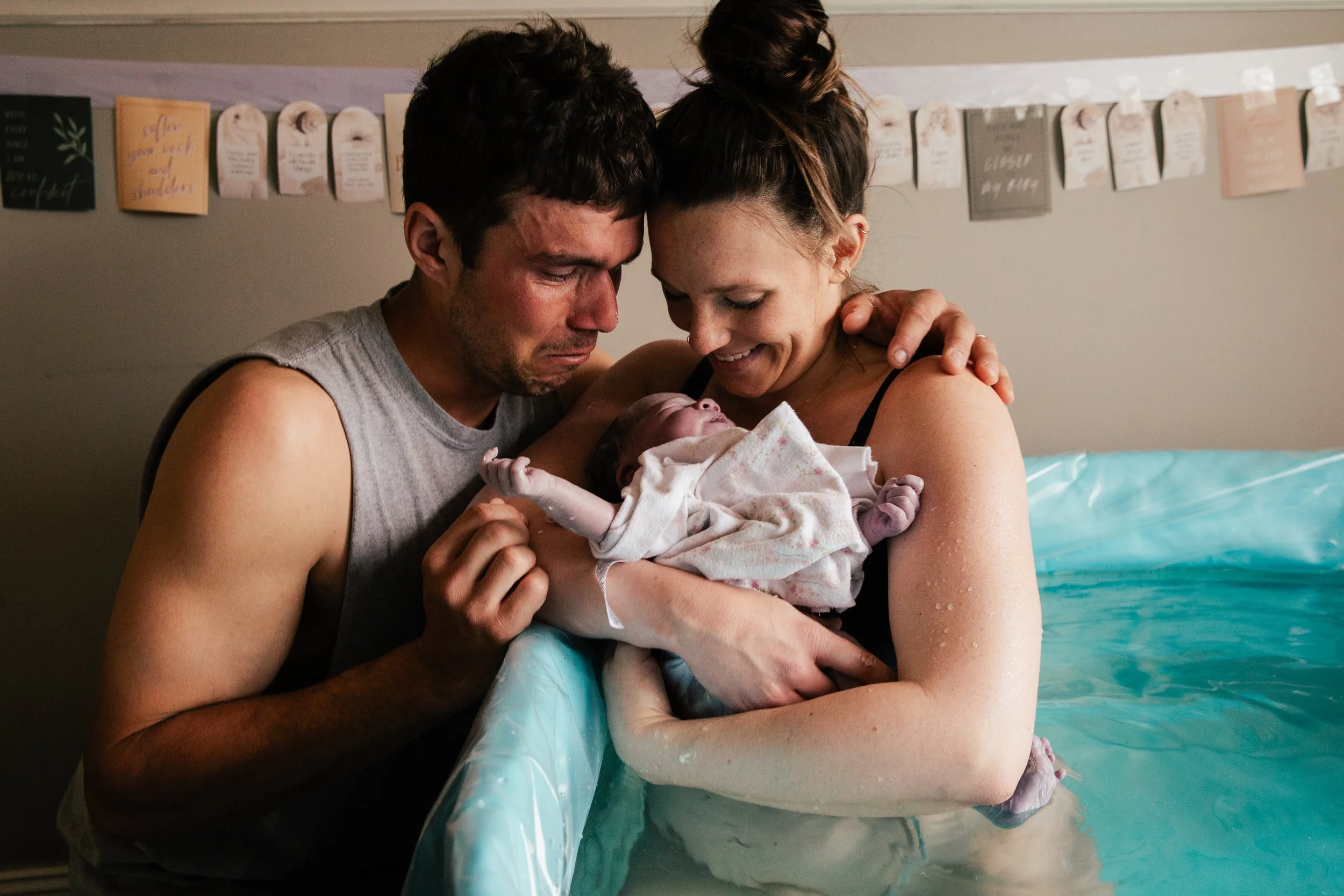 A man and woman holding a newborn baby together in a hospital room, all smiling and looking at the baby affectionately.
