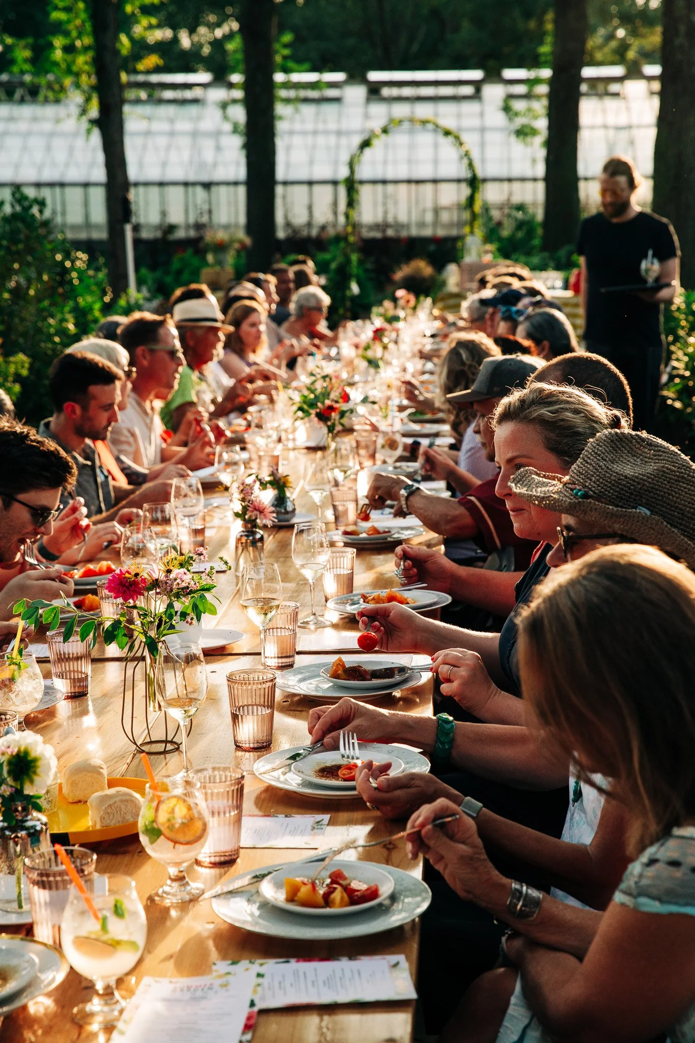 A large group of people gathered around a long outdoor dining table, enjoying a meal in a garden setting during daytime, with green trees and a greenhouse in the background.