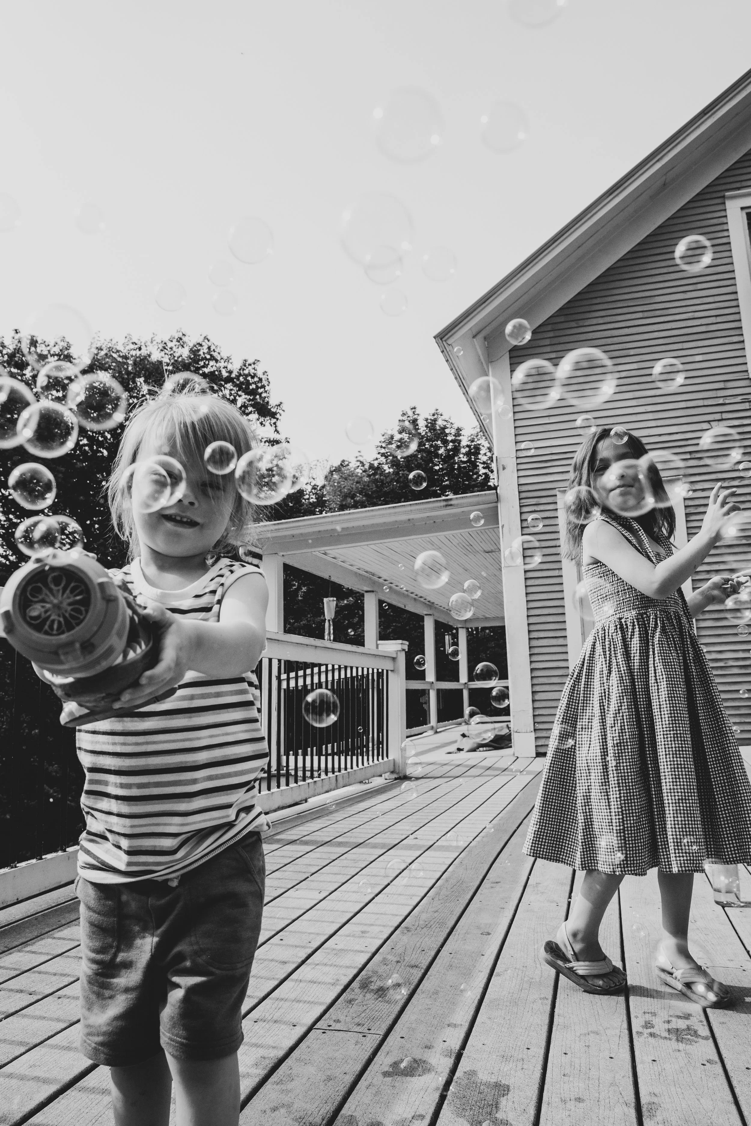 Two young girls playing with bubbles on a wooden deck outside a house. The girl in the foreground has blonde hair covering her face and is holding a bubble gun. The other girl, standing to the right, has dark hair and is wearing a checkered dress. Th