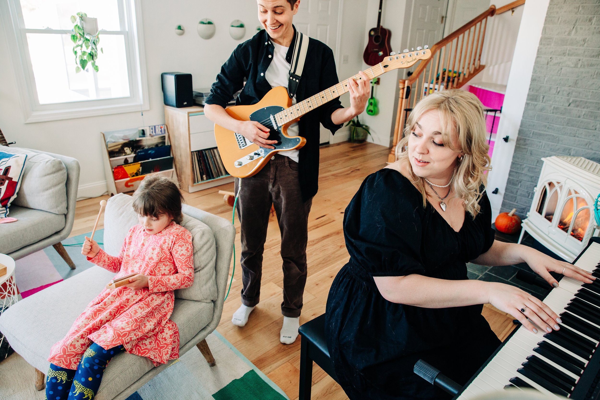 A woman is playing the piano while a young girl sits on a chair with a drumstick in her hand, and a man is playing the guitar. Another woman is sitting nearby, smiling.