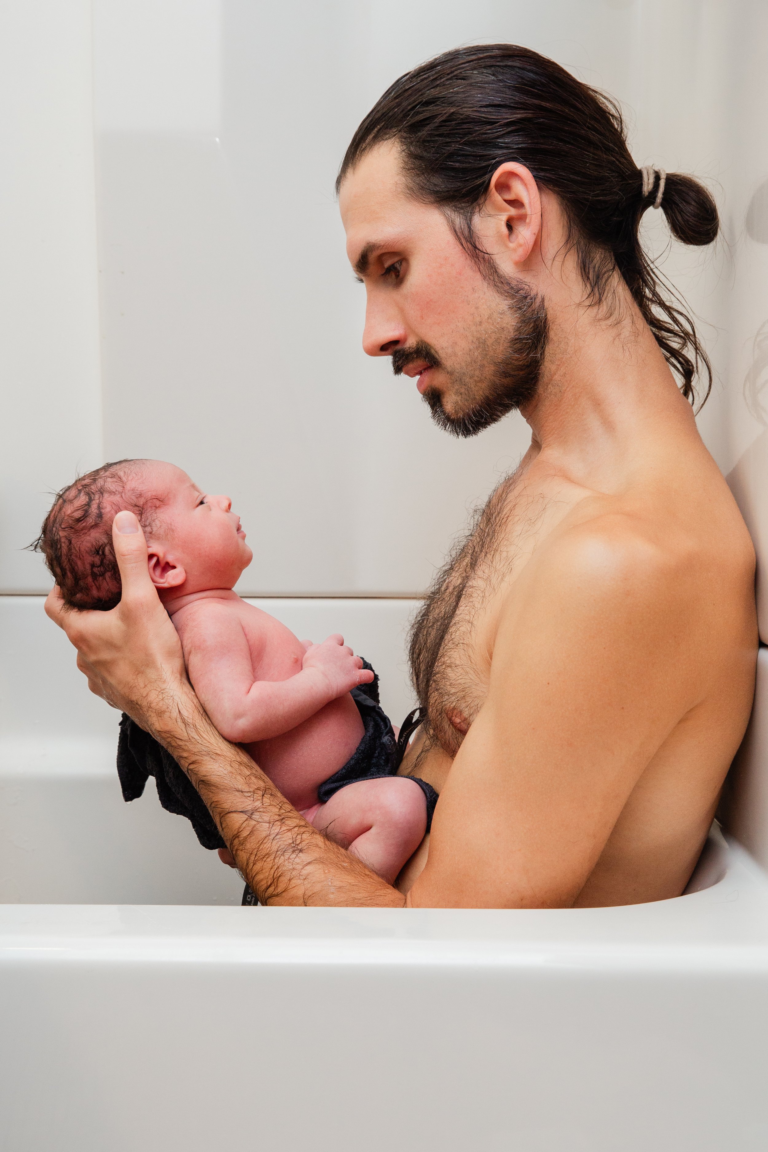 A shirtless man with dark hair and a beard holding a newborn baby with curly hair, in a white bathtub or tub, looking at each other.