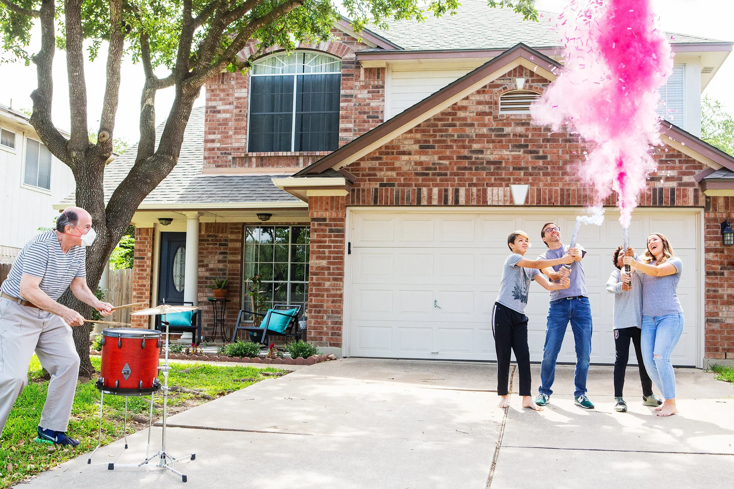 People celebrating with smoke bombs in the driveway of a suburban house, with a drummer playing nearby and family members enjoying the moment.