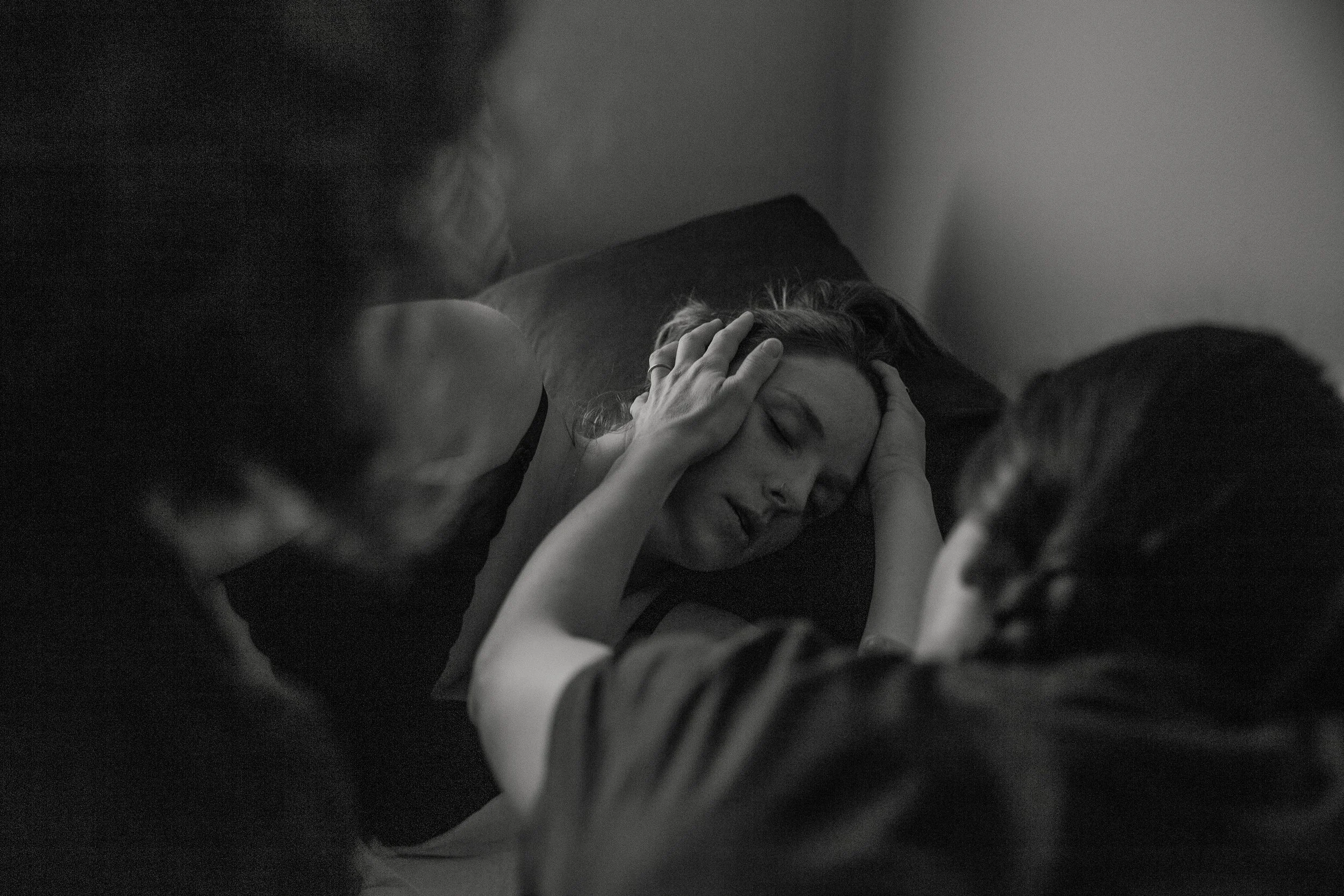 A woman with long hair is laying on a pillow with her eyes closed, holding her head with her hand. Her face shows distress or pain.