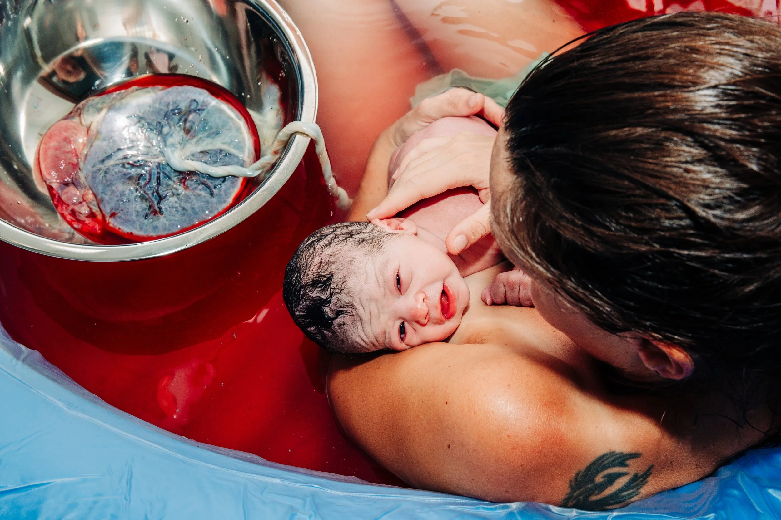 A newborn baby is being held by a woman in a birthing pool during water birth, with the baby's head just emerging from the water and the woman supporting the baby.