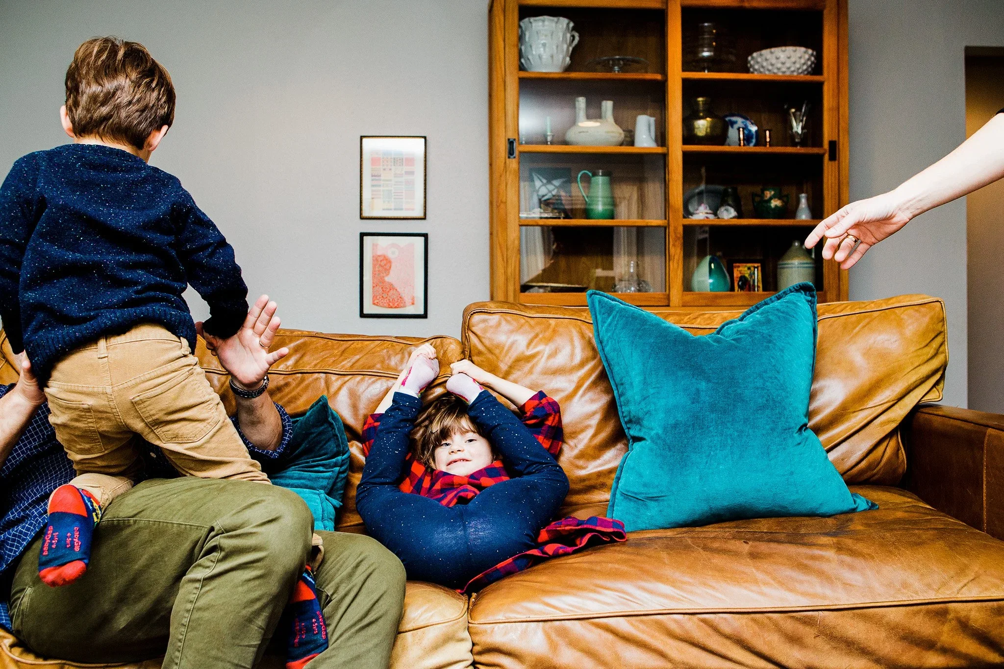 A group of children and an adult playing and relaxing on a tan leather couch in a living room. Some children are lying down, others are sitting or moving around. The scene is lively and casual.