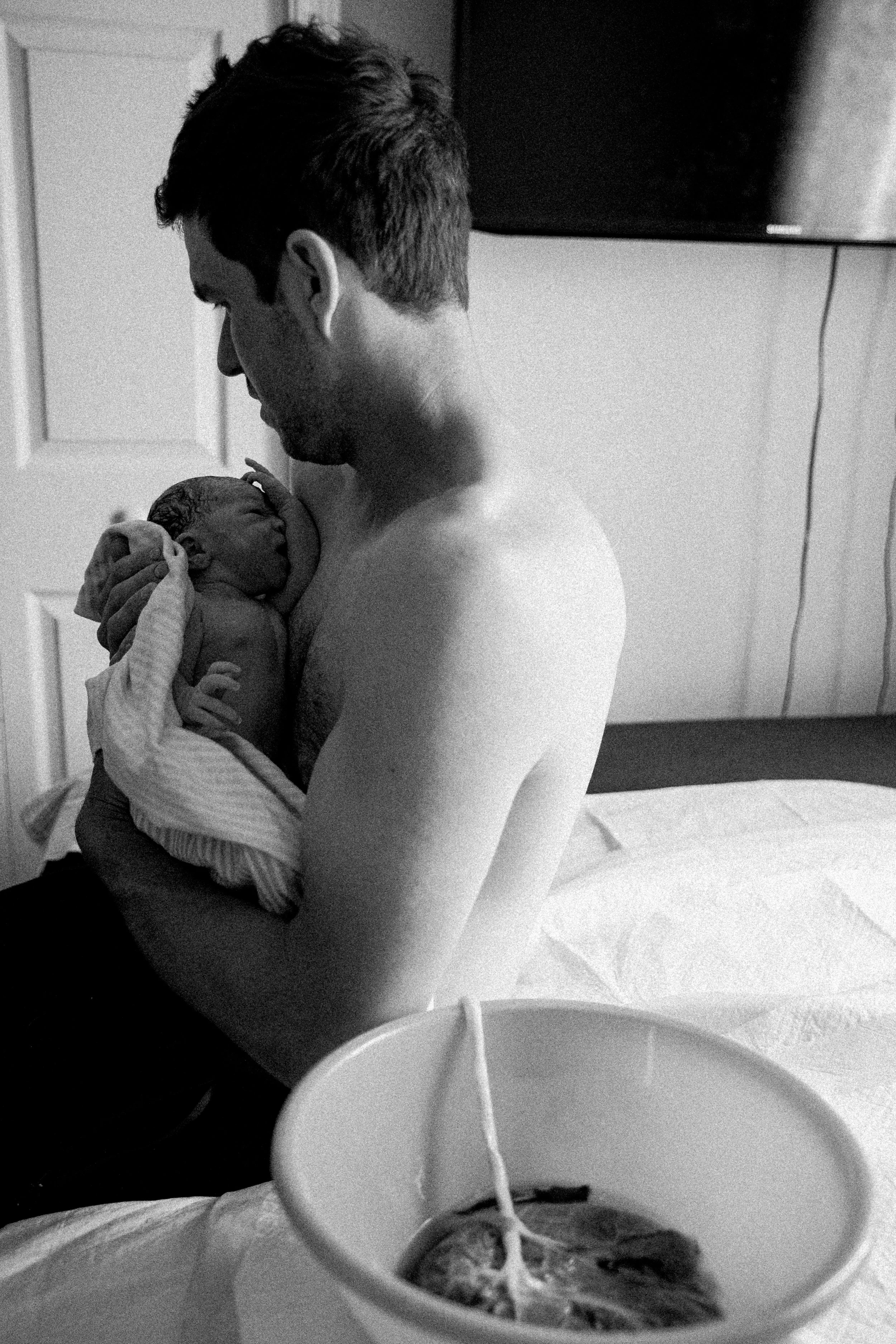 A man holding a newborn in a hospital room, with a clock on the wall, a television screen, and a cup with a stir stick in the foreground.