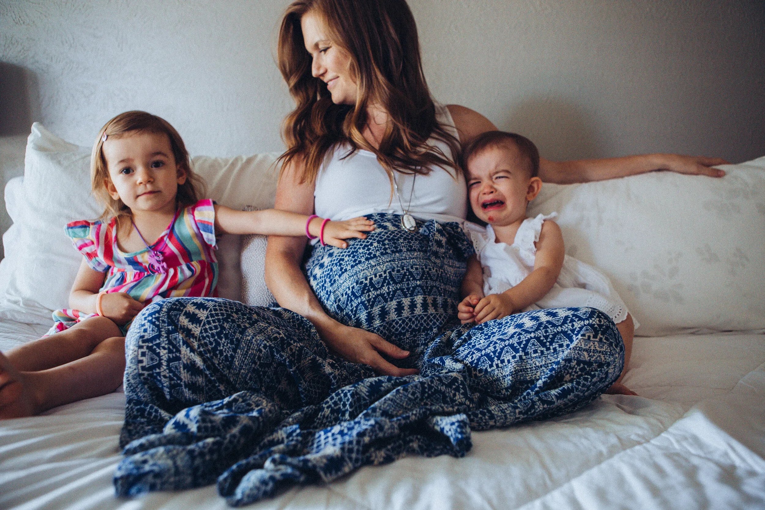 A woman sitting on a bed with two young children, one girl and one boy, in a bedroom with white bedding and a light gray wall. The woman is smiling, the girl appears calm, and the boy is crying.
