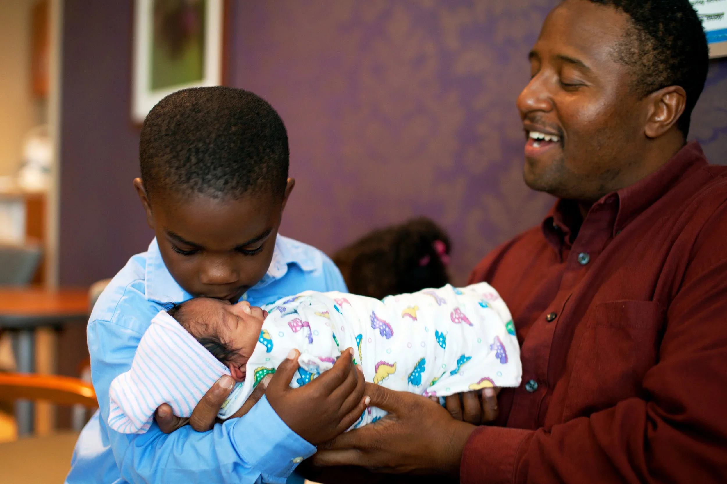 A man and boy caring for a newborn baby in a room with purple walls and framed pictures.