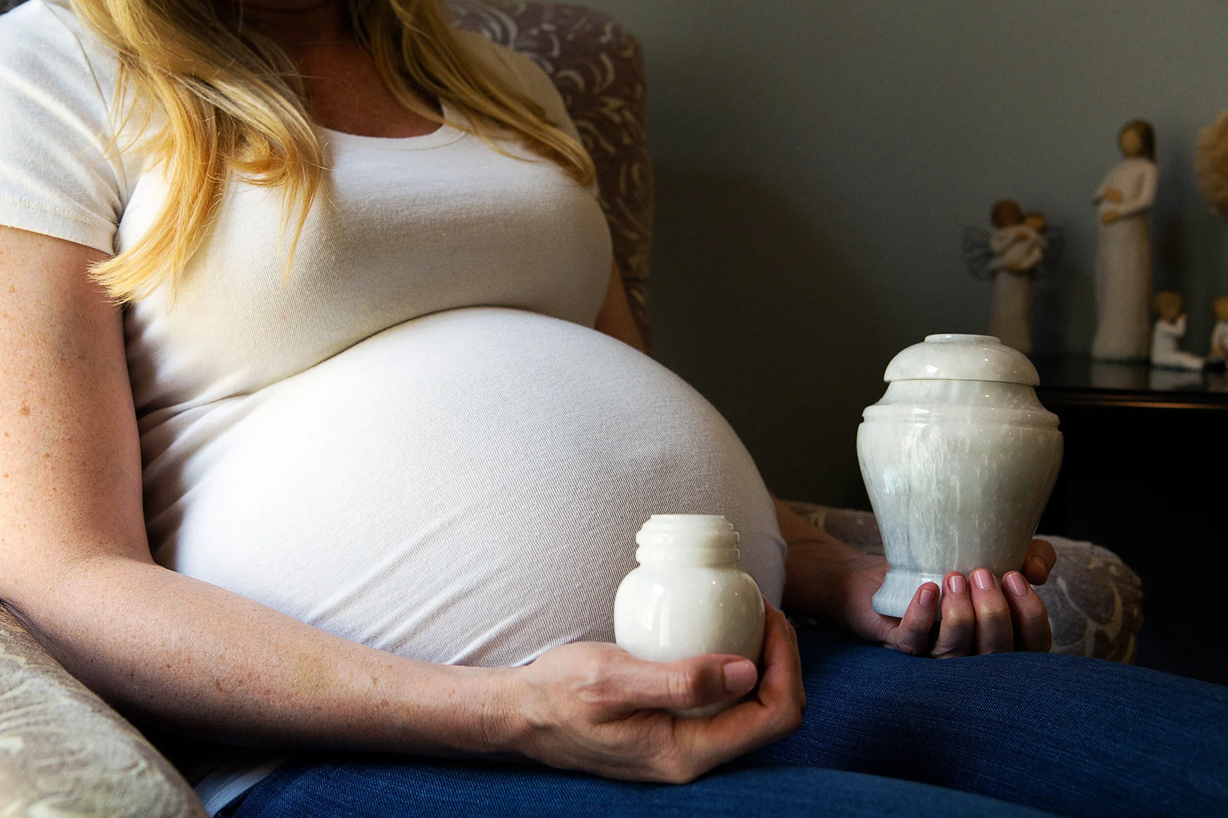 A pregnant woman with blonde hair wearing a white t-shirt sitting on a couch and holding a white ceramic vase.