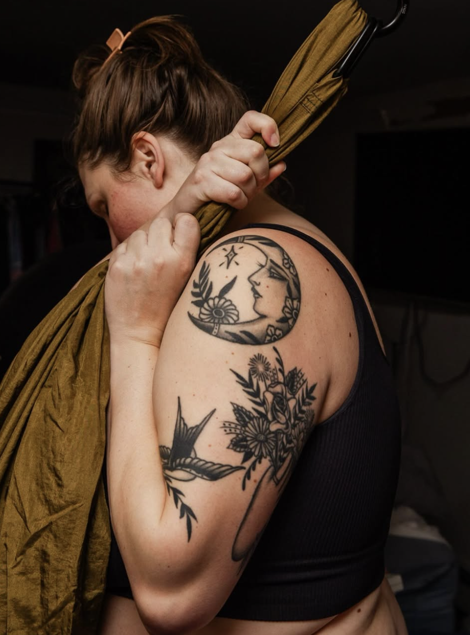 A woman is hugging an umbrella while showing off tattoos on her shoulder and upper arm. The tattoos include a woman's face within a crescent moon and flowers, and a bird among flowers.