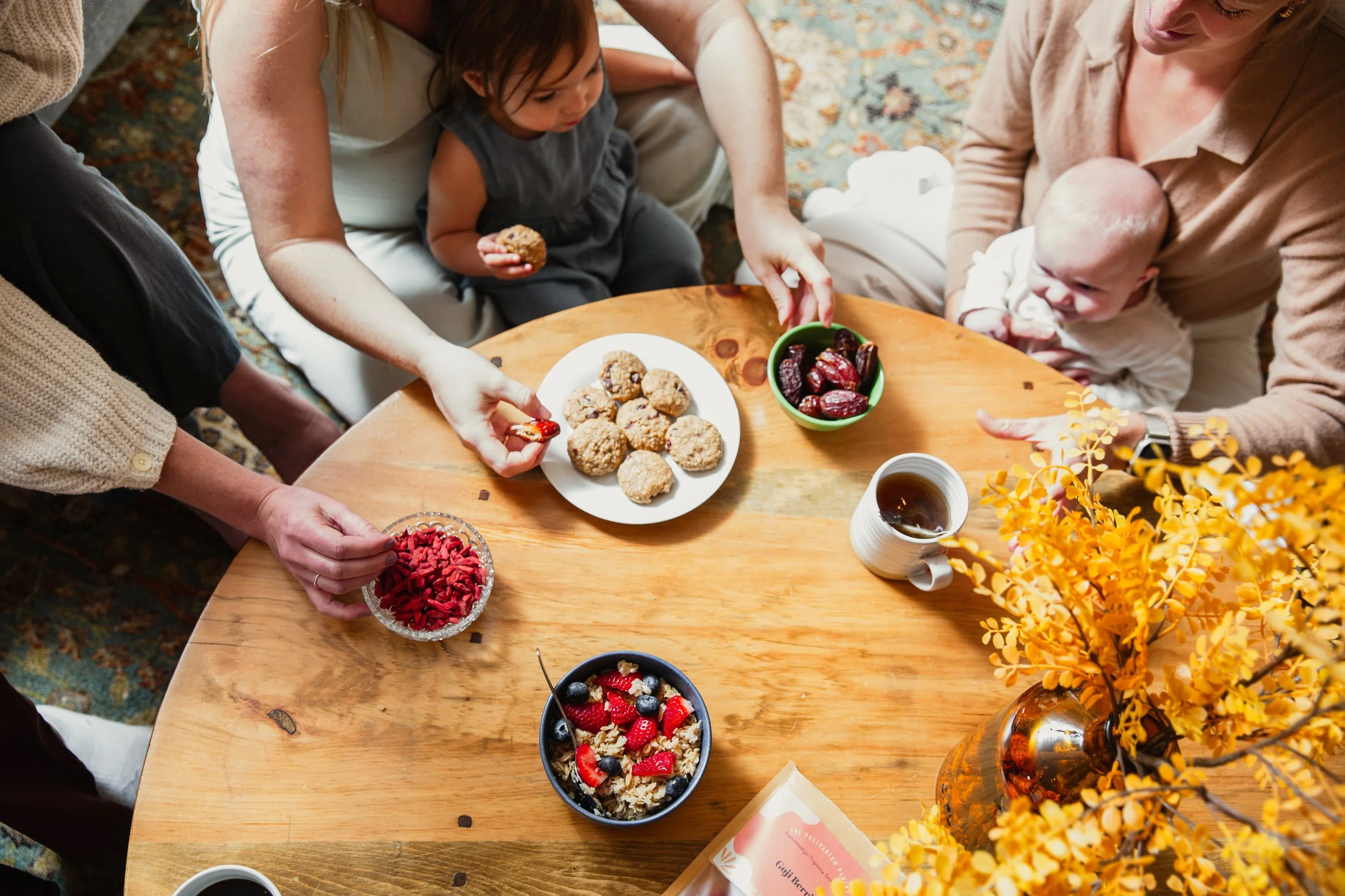 People around a wooden table sharing snacks like cookies, berries, and dates, with a vase of yellow flowers in the foreground.