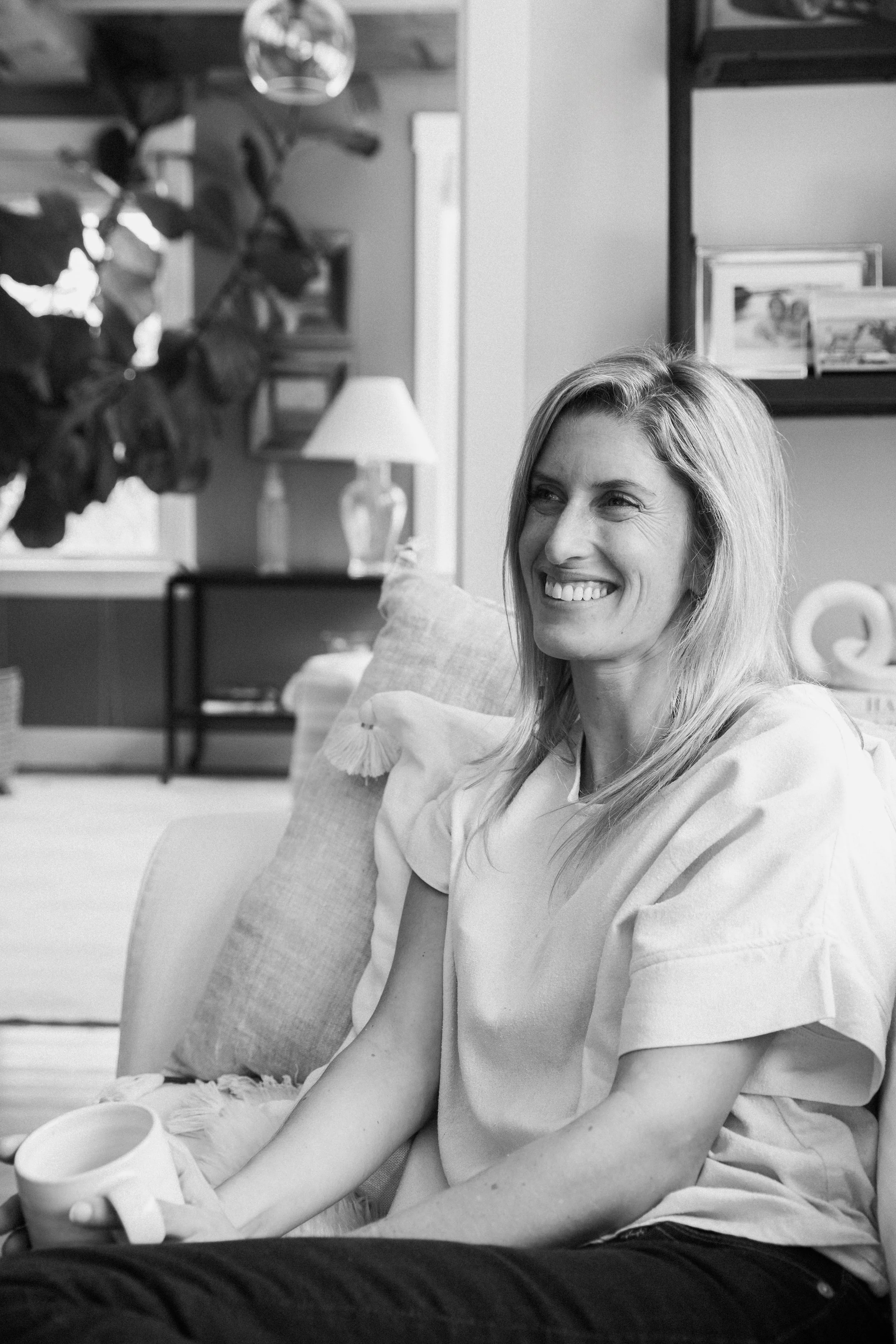A woman with long blonde hair smiling while sitting on a couch at home, holding a mug, with a lamp, picture frames, and a shelf in the background.