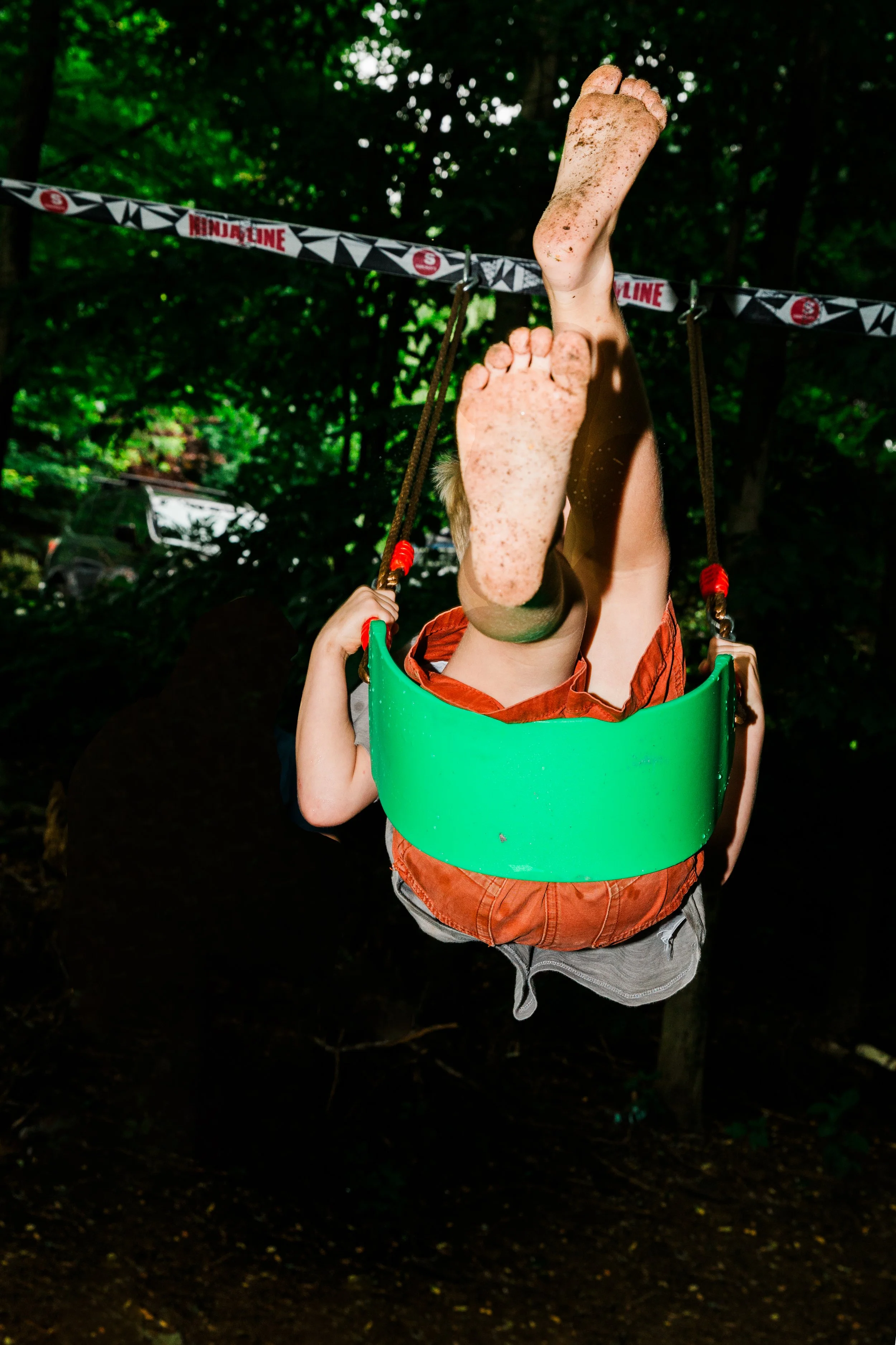 Child swinging upside down on a playground swing with dirty feet and legs, wearing orange shorts, with caution tape in the background.