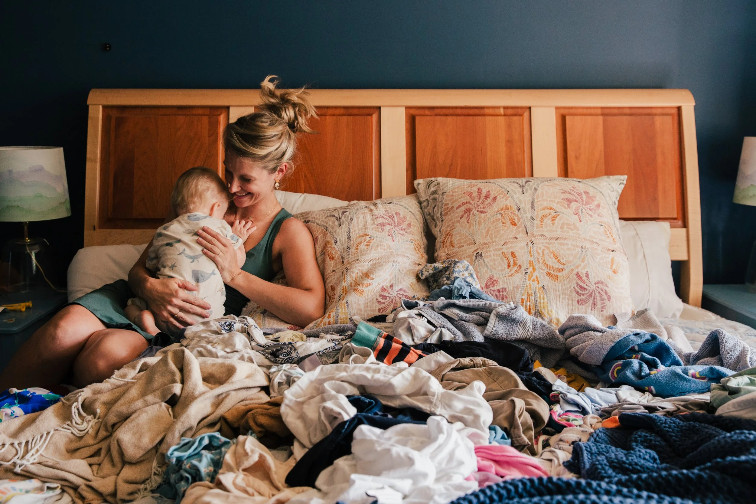 A woman and a young child sitting on a bed surrounded by piles of laundry, sharing a moment of joy and affection.