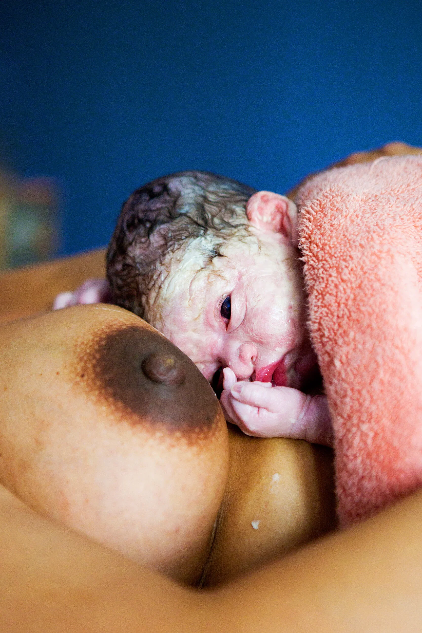 A newborn baby with closed eyes, pinkish skin, and wet hair, resting on a person's chest, holding their finger, with baby's head partly covered with a peach-colored towel.