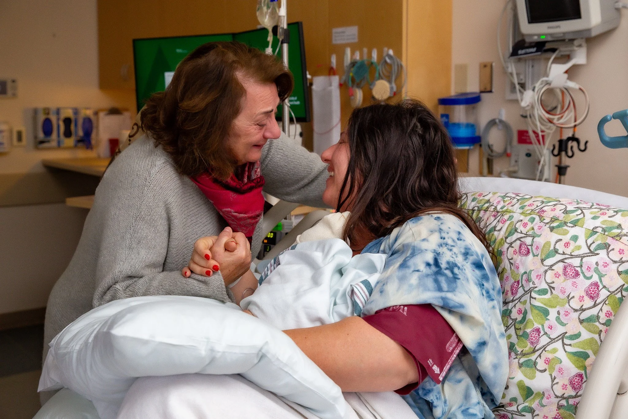 A woman and a girl in a hospital room are smiling and holding hands, with the woman leaning over the hospital bed to be face-to-face with the girl.