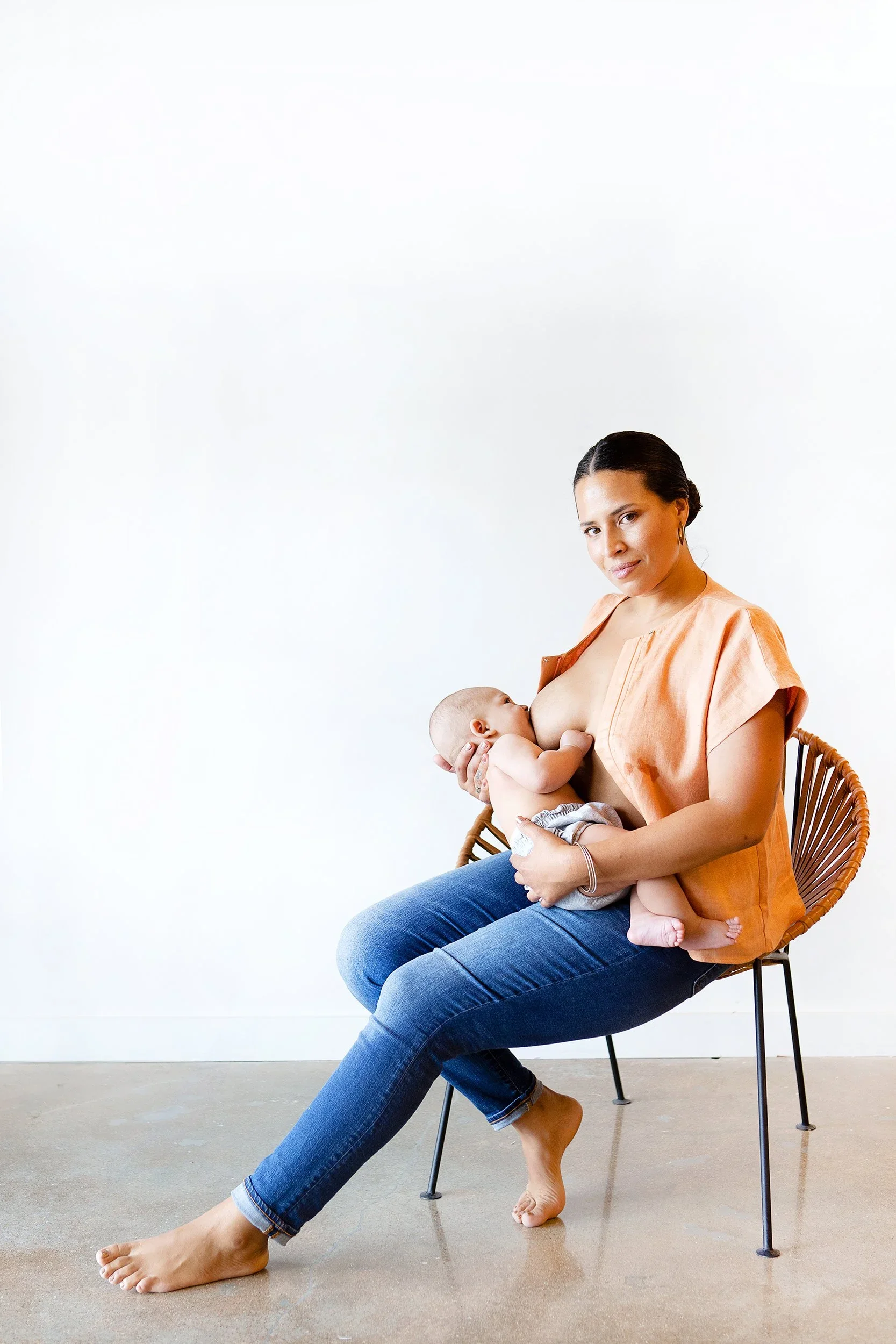 A woman sitting in a chair, breastfeeding a baby. She is wearing a peach-colored top and blue jeans, with a calm expression. The background is white and minimalistic.
