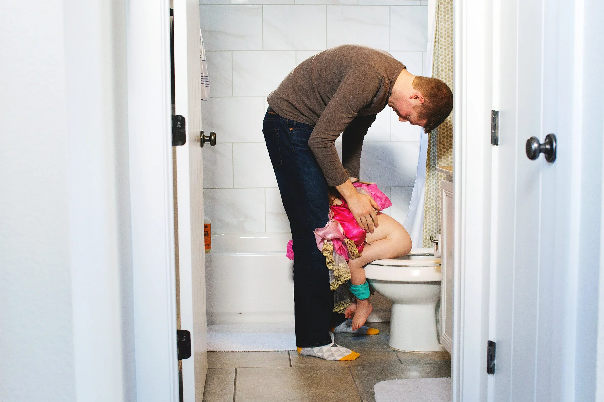 A man leaning over a young girl sitting on a toilet seat inside a bathroom. The girl appears to be distressed, and the man is holding her arm as if trying to comfort her. The bathroom has white tiles and partially open doors.