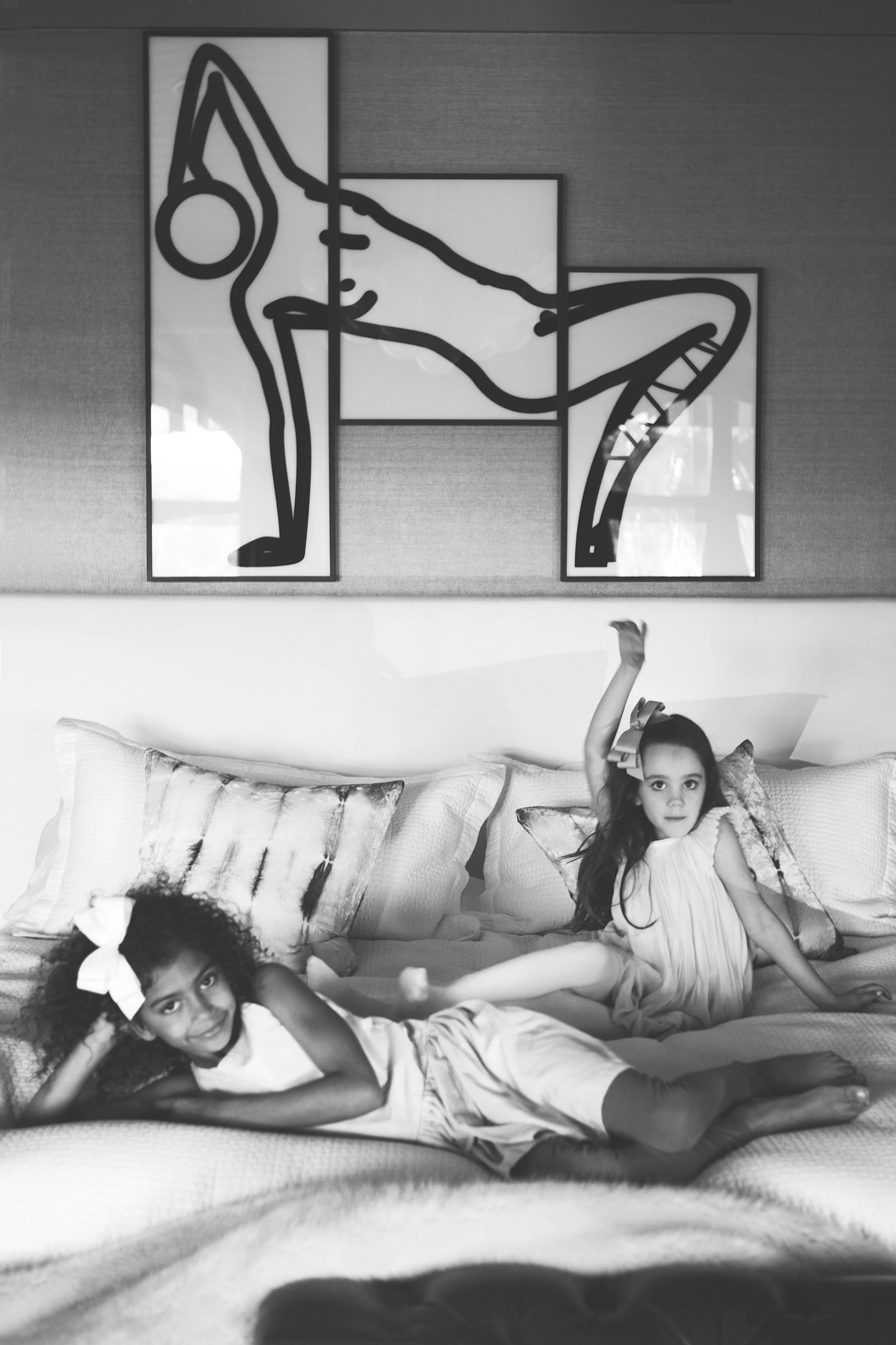 Two young girls lying on a bed with decorative pillows, raising their arms. One girl with curly hair and a large bow is in the foreground, and another girl with straight hair is in the background, both looking at the camera. A large abstract art piec
