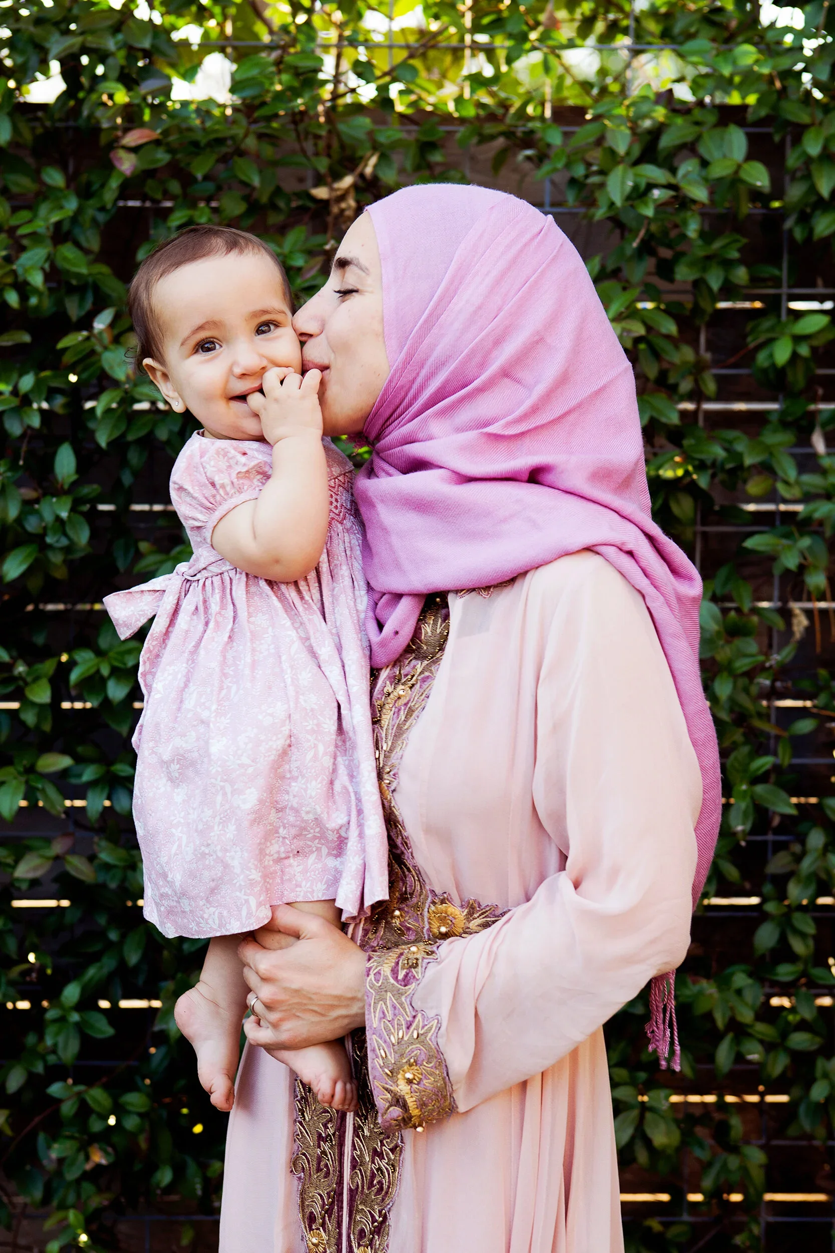 A woman wearing a pink hijab holding a smiling young girl in a pink dress, outdoors with green leafy bushes in the background