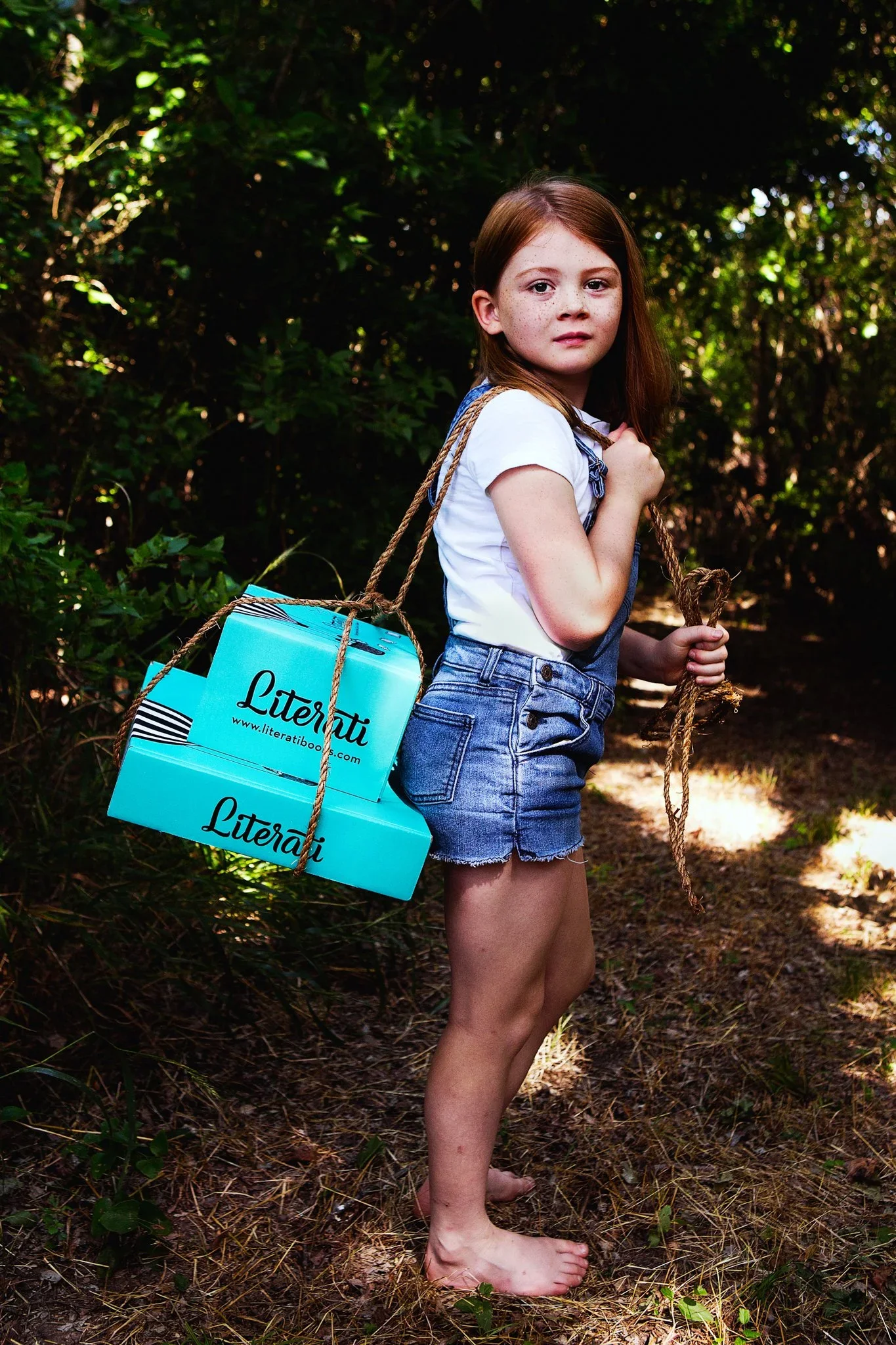 A young girl with red hair and freckles stands barefoot in a forest, holding a rope over her shoulder with a blue box labeled Literati hanging from it. She wears a white t-shirt and denim shorts, and looks at the camera with a neutral expression.