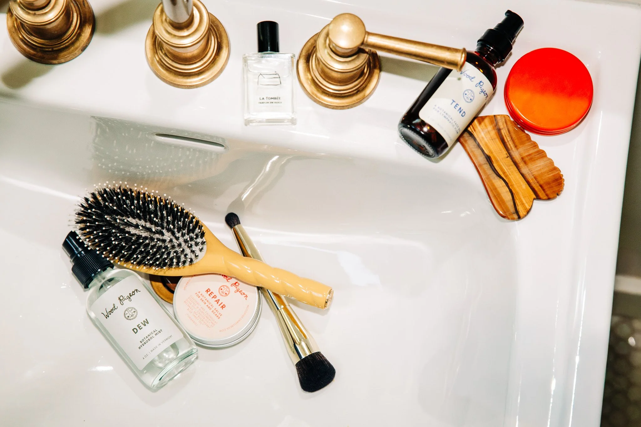 A collection of personal care and grooming items including a hairbrush, a small bottle of perfume labeled 'Wood Fig fuel,' a bottle of hydrosol mist labeled 'Dew,' a round tin labeled 'Repair,' a makeup brush, and a few candles with golden holders.
