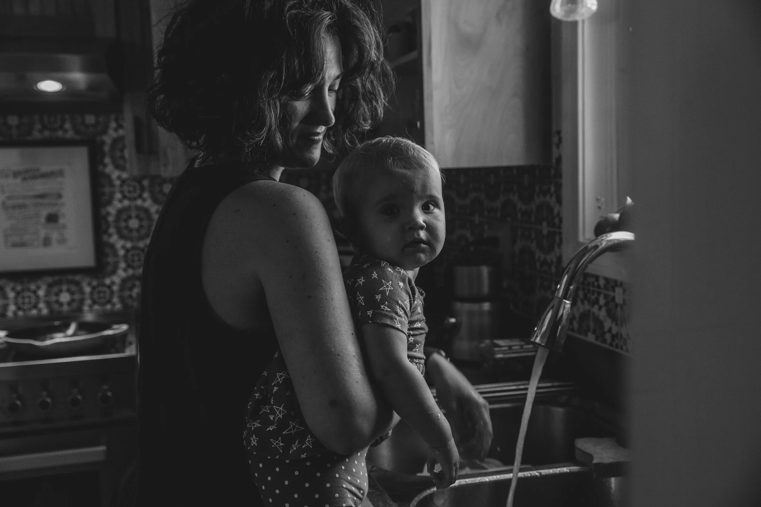 A woman with curly hair holds a young child while washing dishes at a kitchen sink.